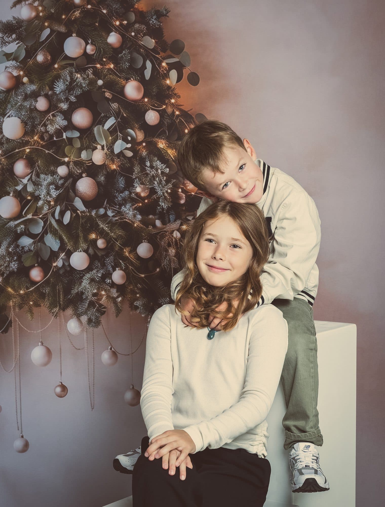 Un frère et sa sœur en tons clairs devant le grand sapin de Noël du studio, décoré de boules
