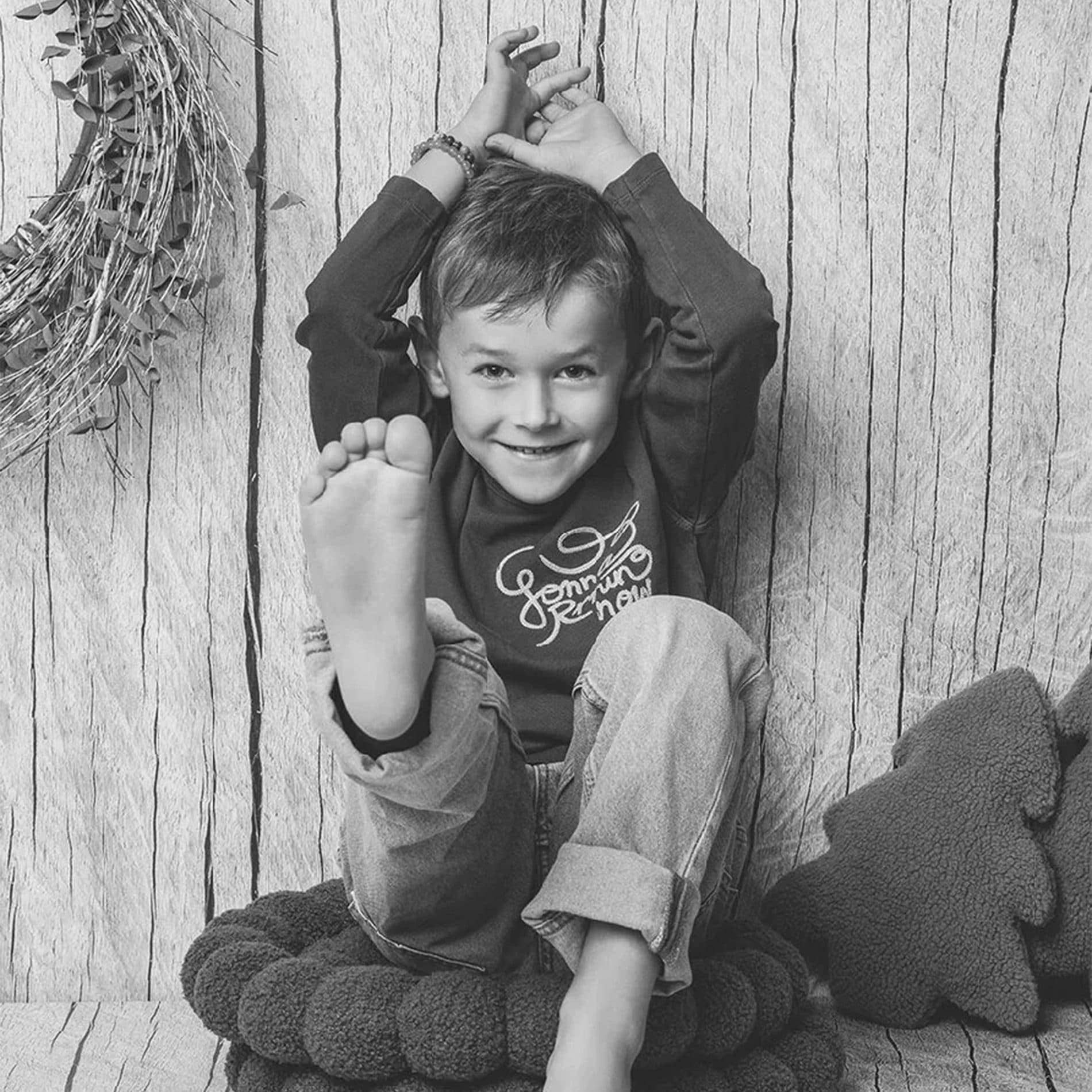 Un petit garçon rieur assis sur un coussin tressé, un pied levé, en noir et blanc