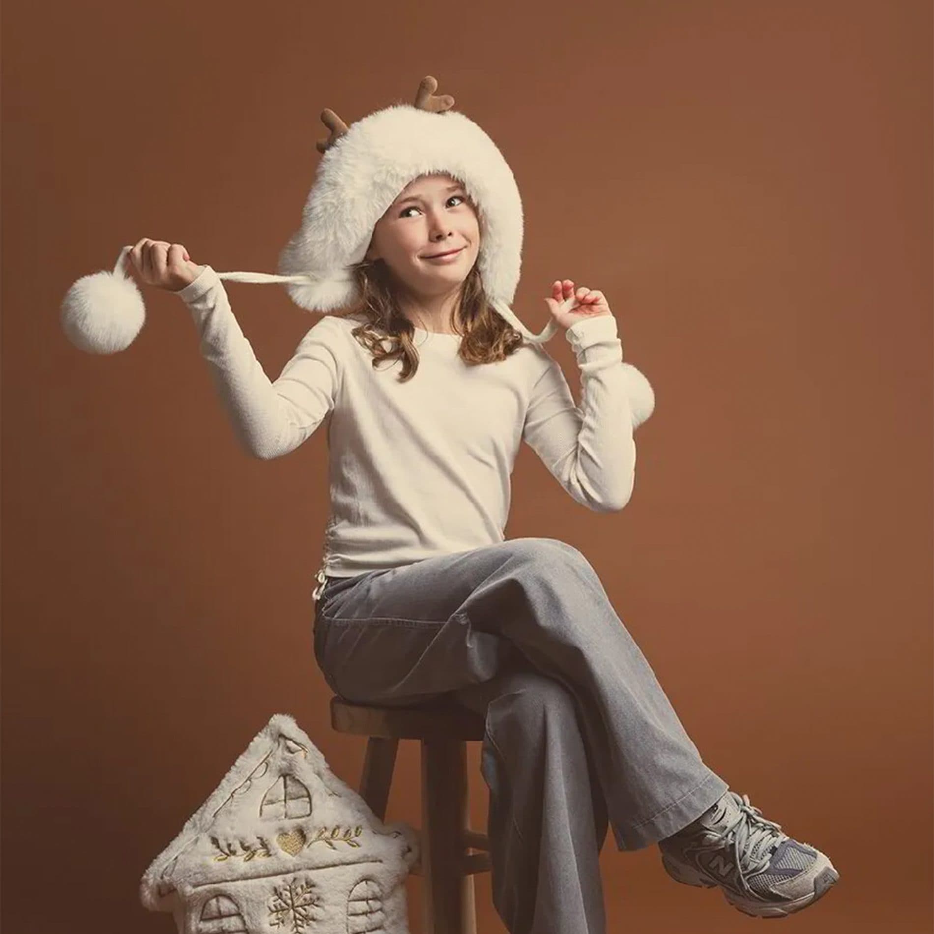 Une fillette en bonnet blanc à pompon assise sur un tabouret, devant un fond brun cacao