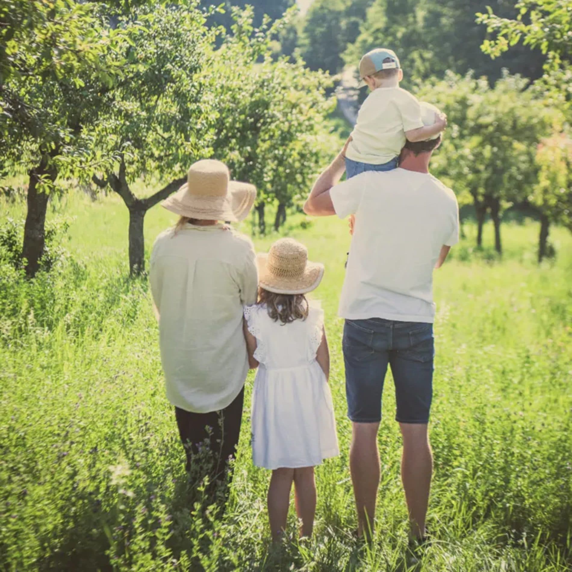 Une famille de dos dans un verger, le papa porte son enfant sur les épaules