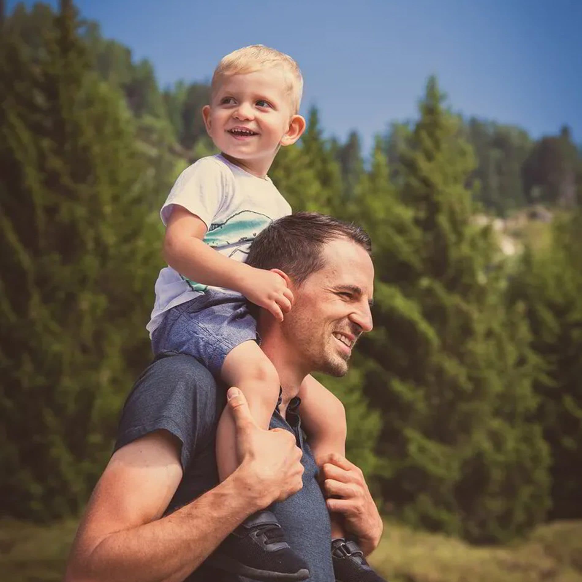 Un papa porte son petit garçon rieur sur les épaules, devant des sapins