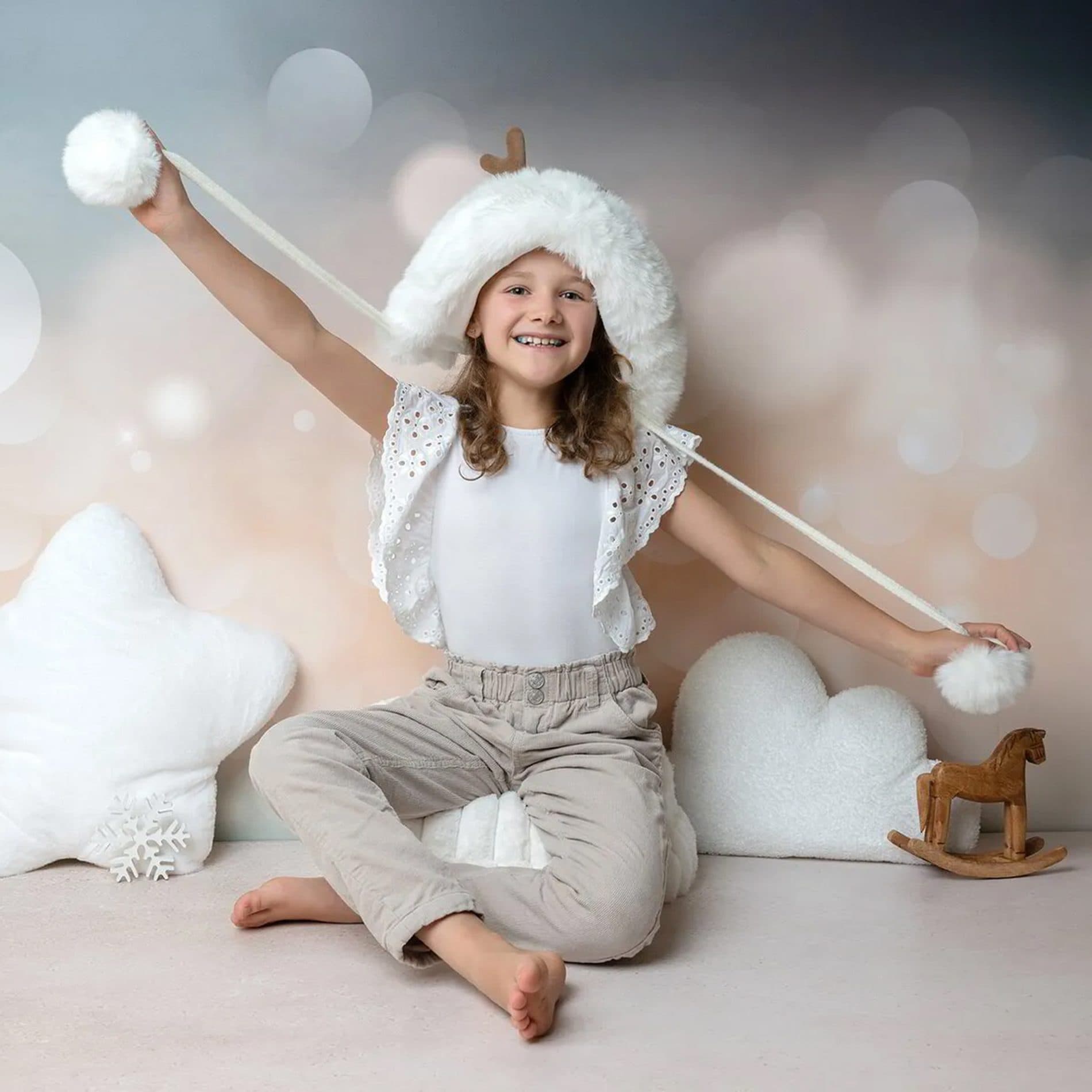 Une fillette en bonnet blanc à pompon assise parmi des coussins en forme de nuage et d’étoile