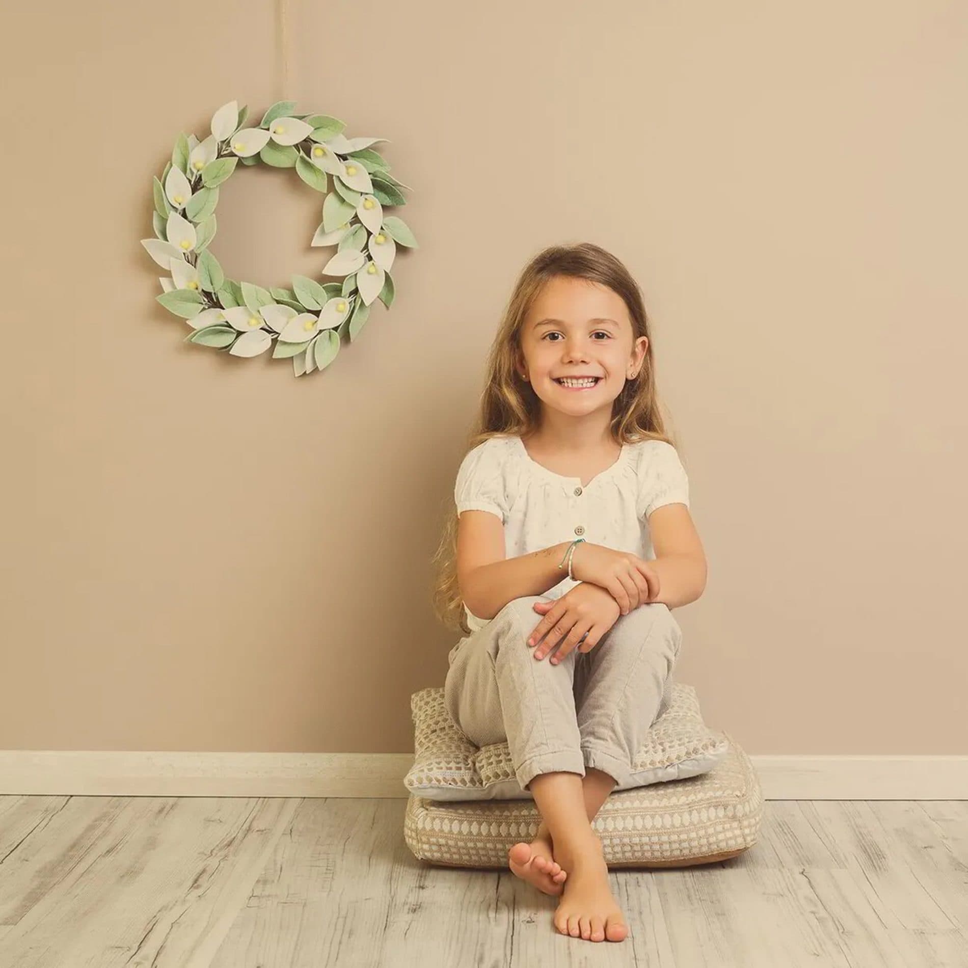 Une fillette assise sur des coussins sous une couronne de verdure, devant un fond beige