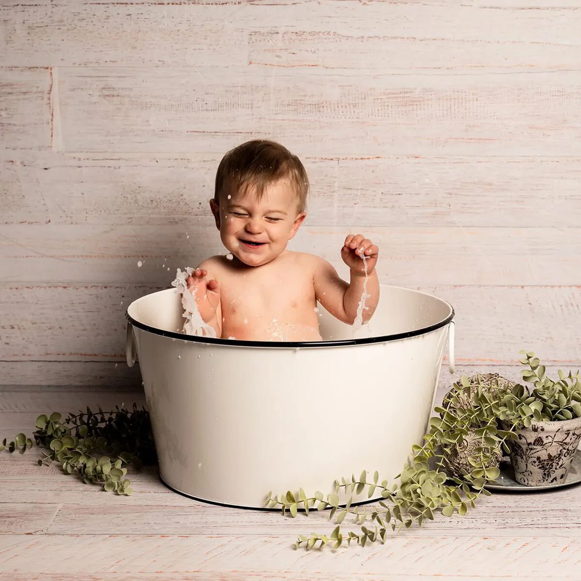 Un bébé rit dans une bassine émaillée, entouré de feuillage
