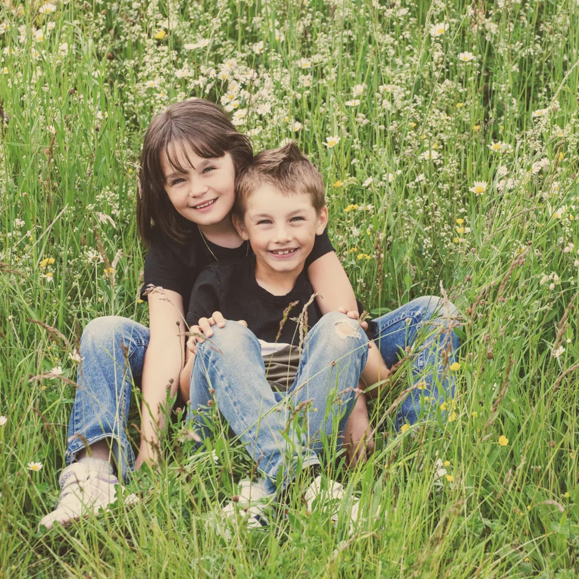Un frère et sa sœur en jean et t-shirts foncés, assis dans les hautes herbes