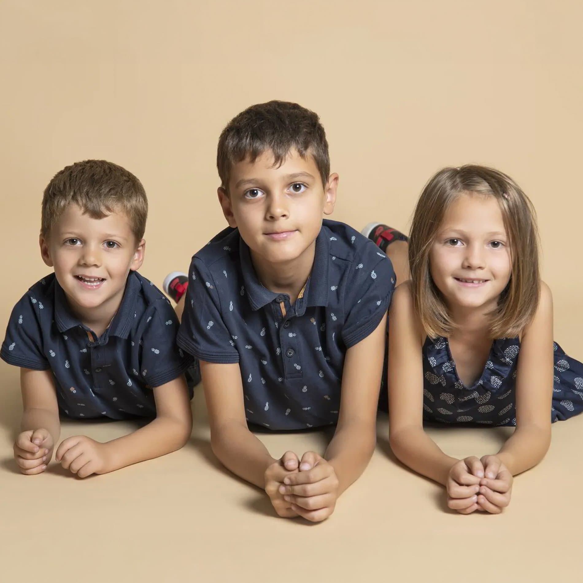 Trois enfants allongés sur le ventre, les mains jointes, sur fond beige