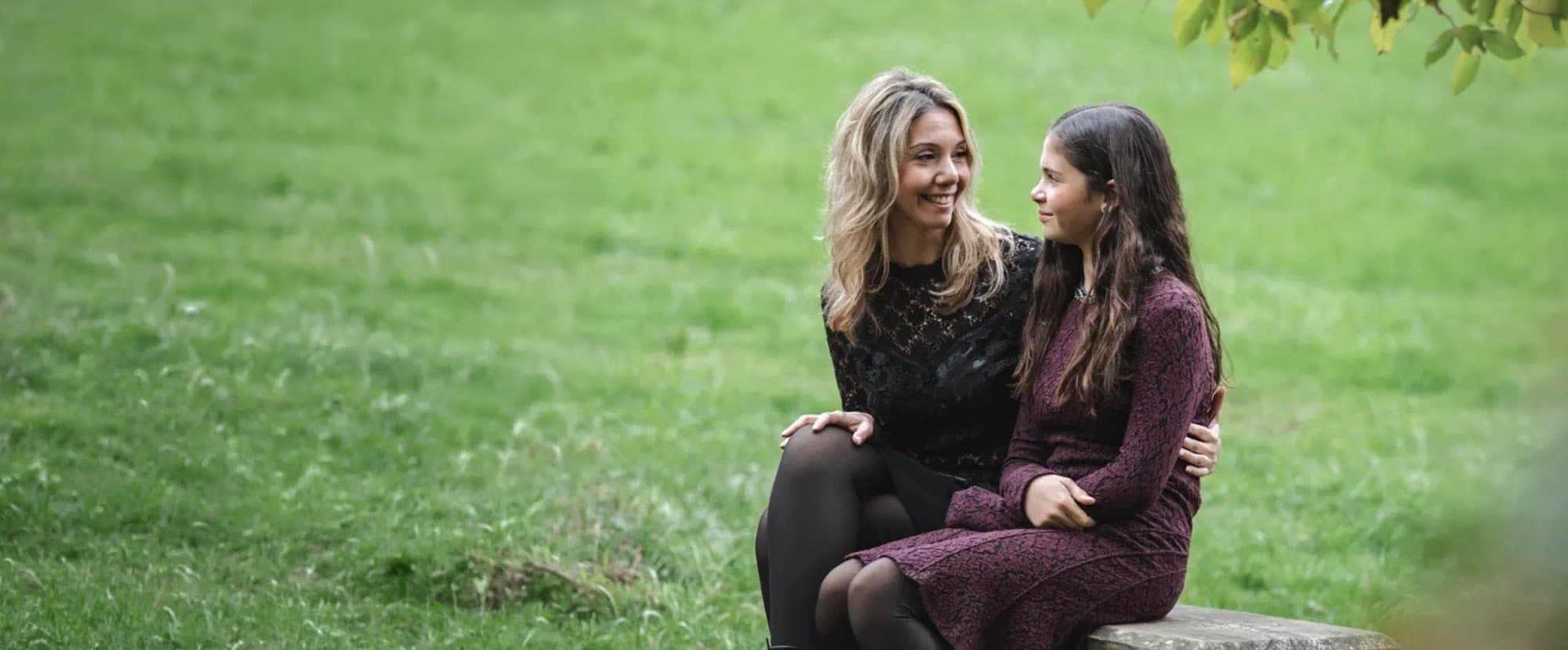 Une maman et sa fille assises sur un banc de pierre dans l’herbe, qui se regardent en souriant