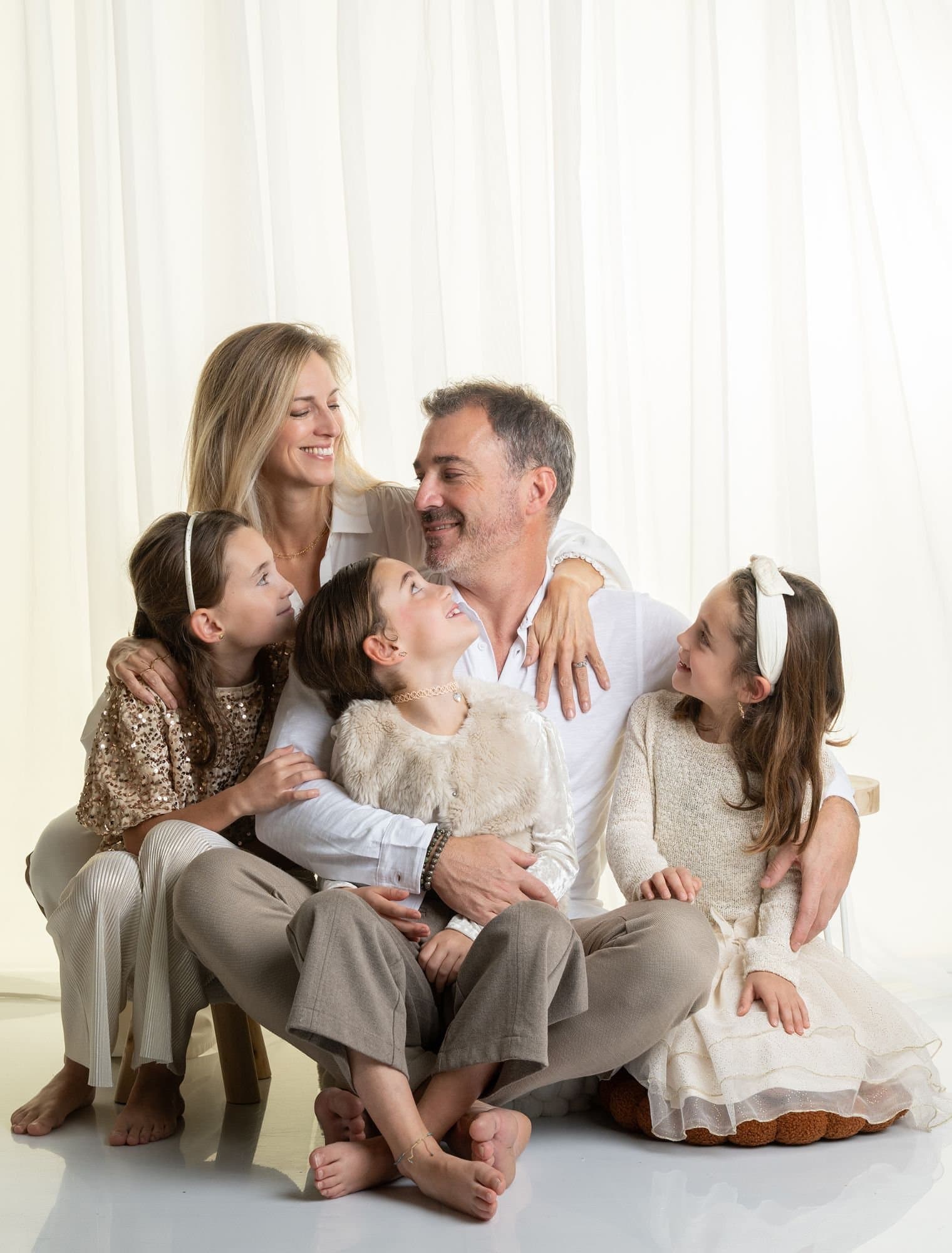 Des parents assis par terre avec leurs trois filles qui les regardent en riant, en tons blancs et beiges devant un rideau clair