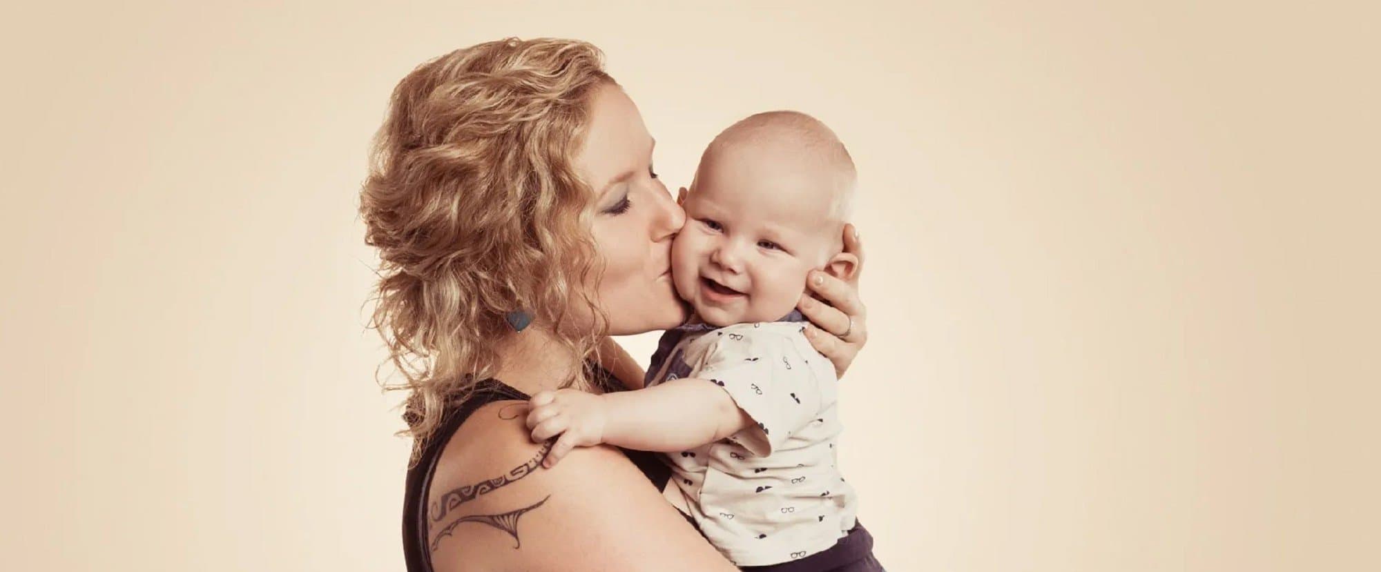 Une maman embrasse la joue de son bébé souriant, sur fond beige