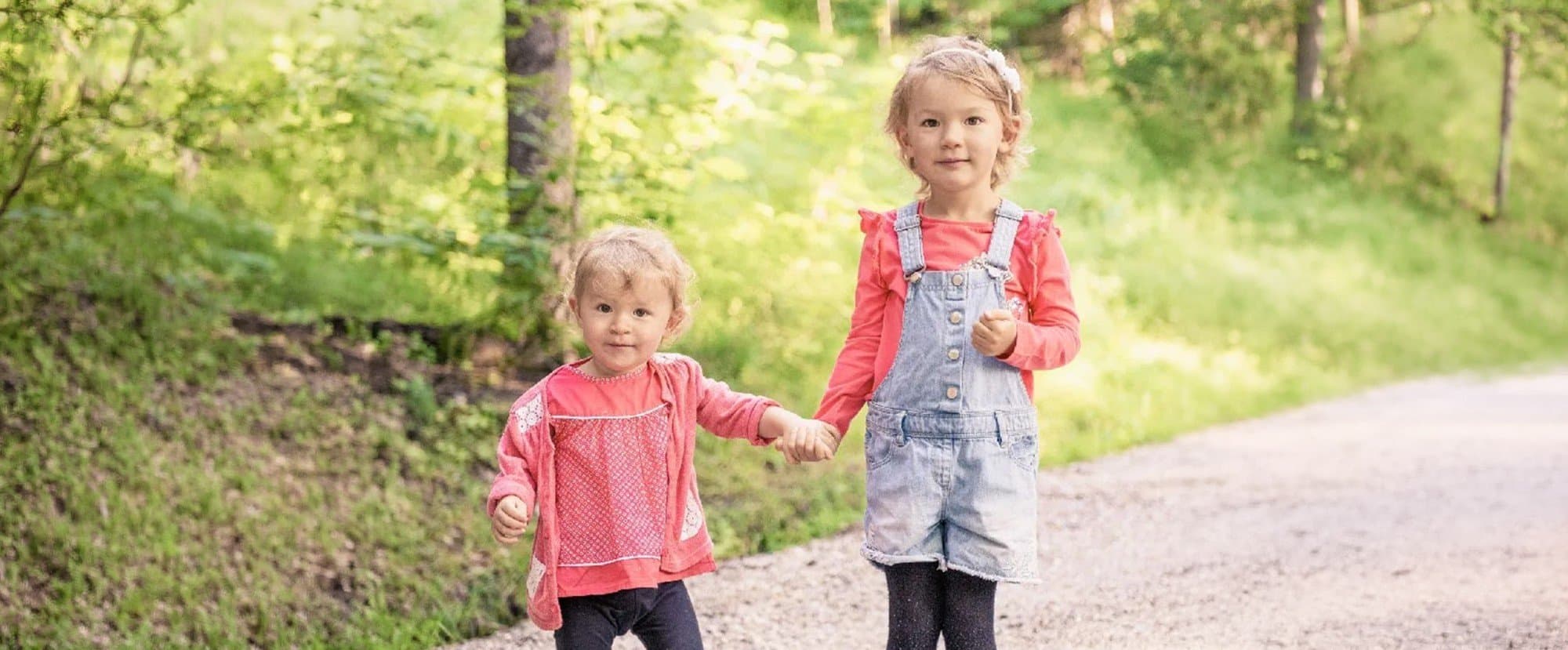 Deux petites sœurs main dans la main sur un chemin en forêt, en tenues corail