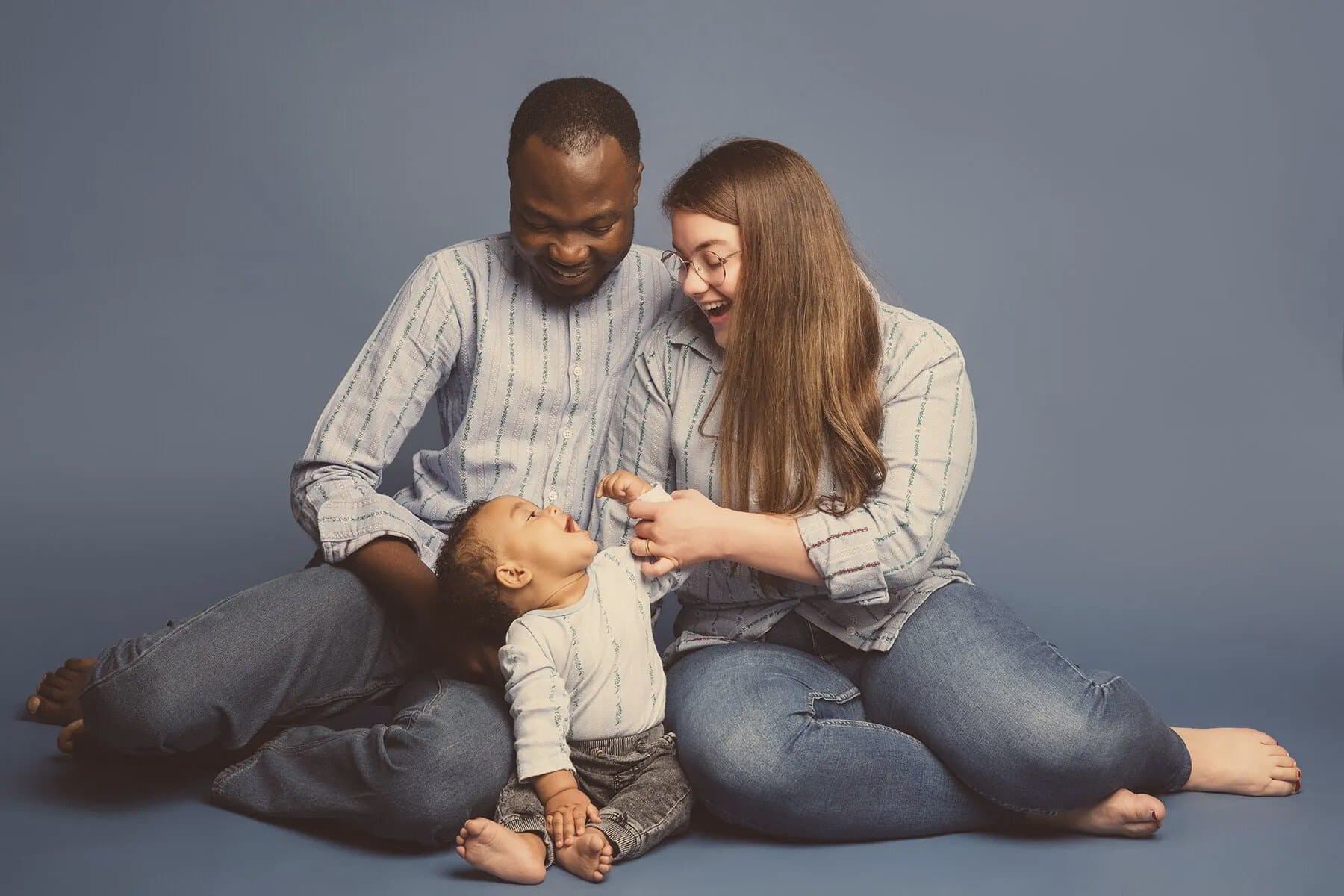 Des parents en chemises claires assorties et en jean rient avec leur bébé assis devant eux, sur fond bleu