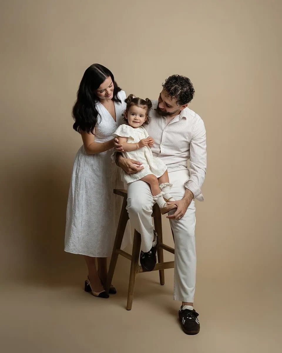 Des parents en blanc et crème avec leur fillette assise sur un tabouret en bois, sur fond beige