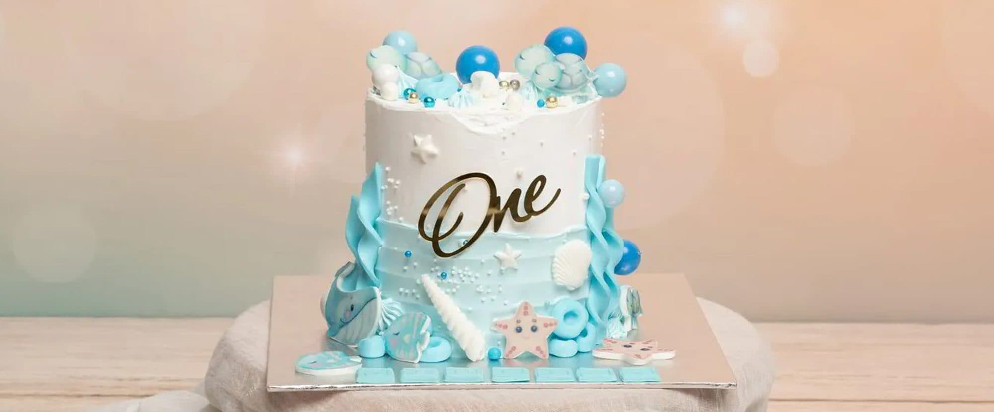 Un gâteau d’anniversaire blanc et bleu décoré de coquillages et d’un « One » doré, sur un fond pêche