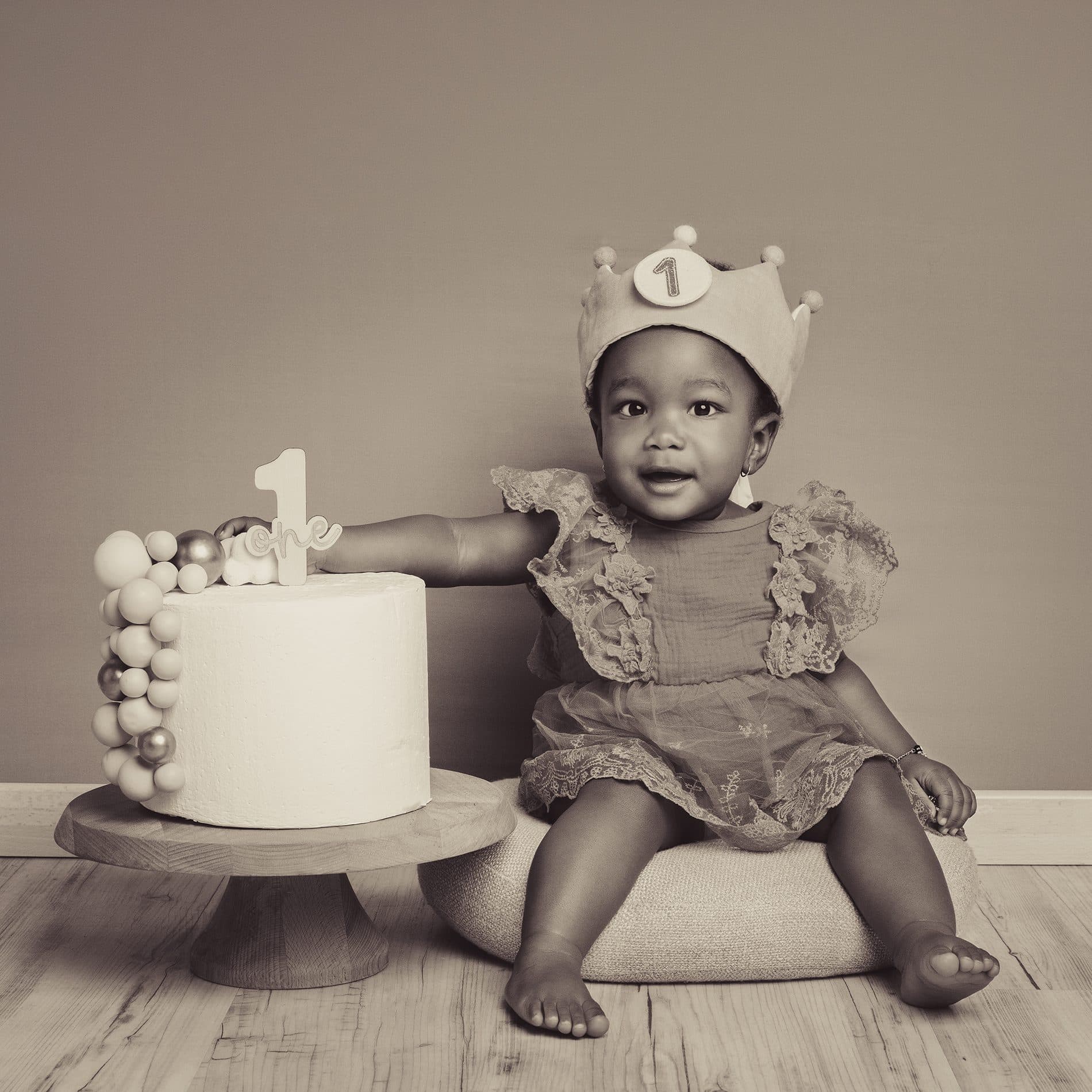 Une fillette coiffée d’une couronne « 1 » assise à côté de son gâteau d’anniversaire blanc, en tons sépia