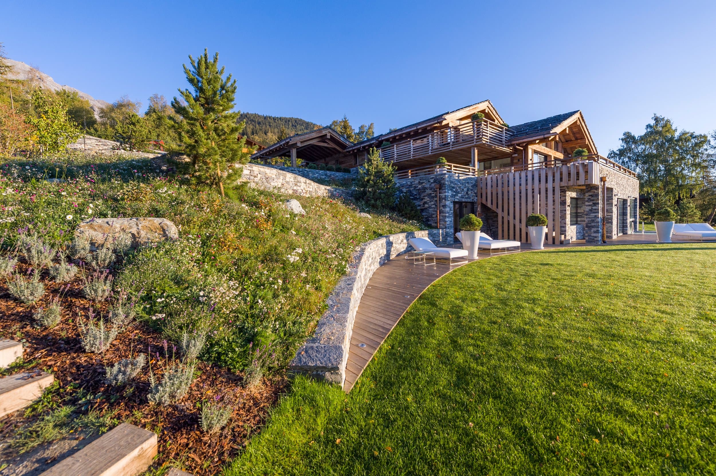 Chalet en bois et en pierre au-dessus d’une pelouse en courbe et d’un talus de rocaille fleuri, jardin aménagé par Arbora