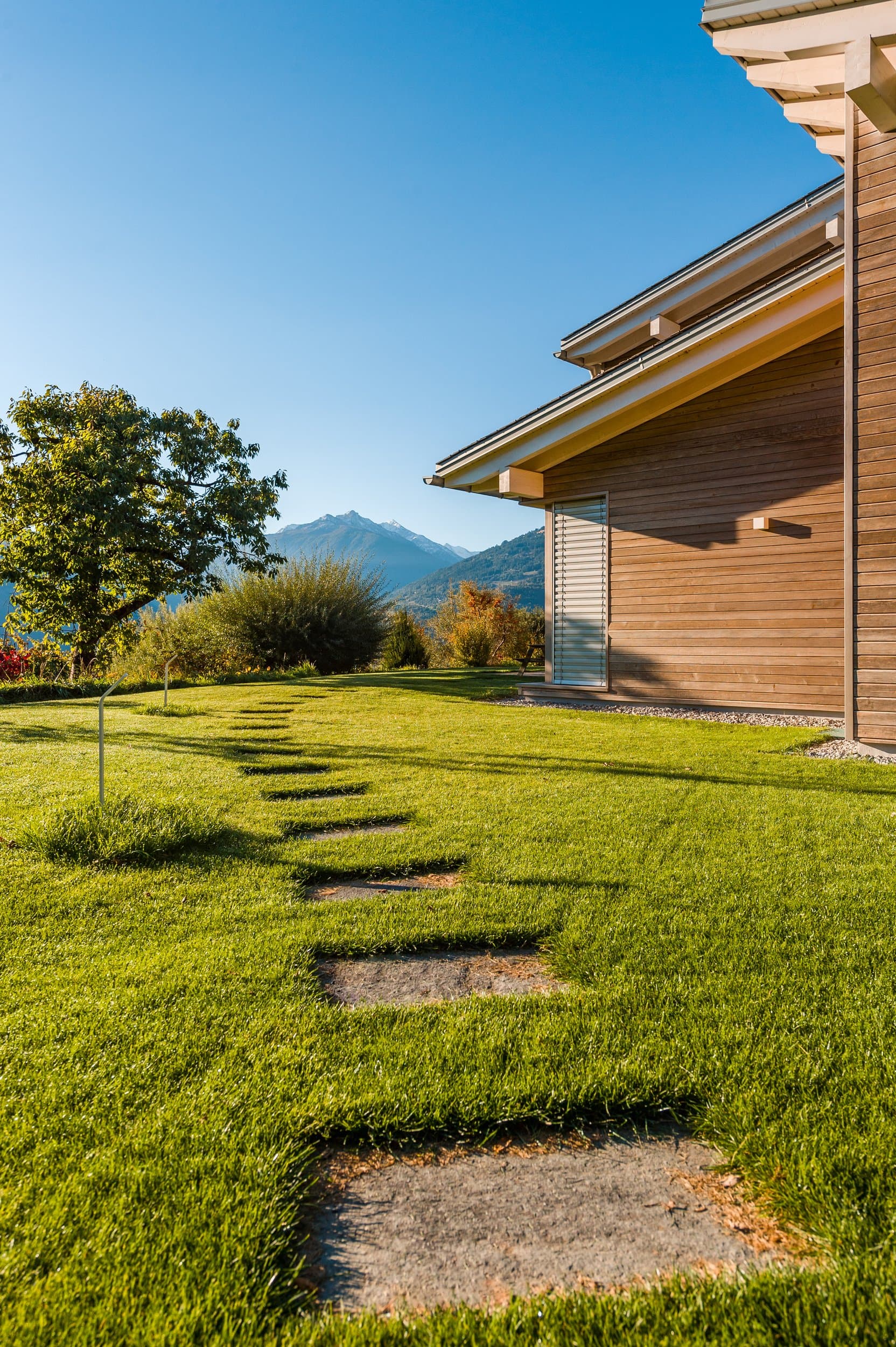 Pas japonais dans une pelouse, jusqu’à une maison au bardage en bois, un arbre et les montagnes au fond