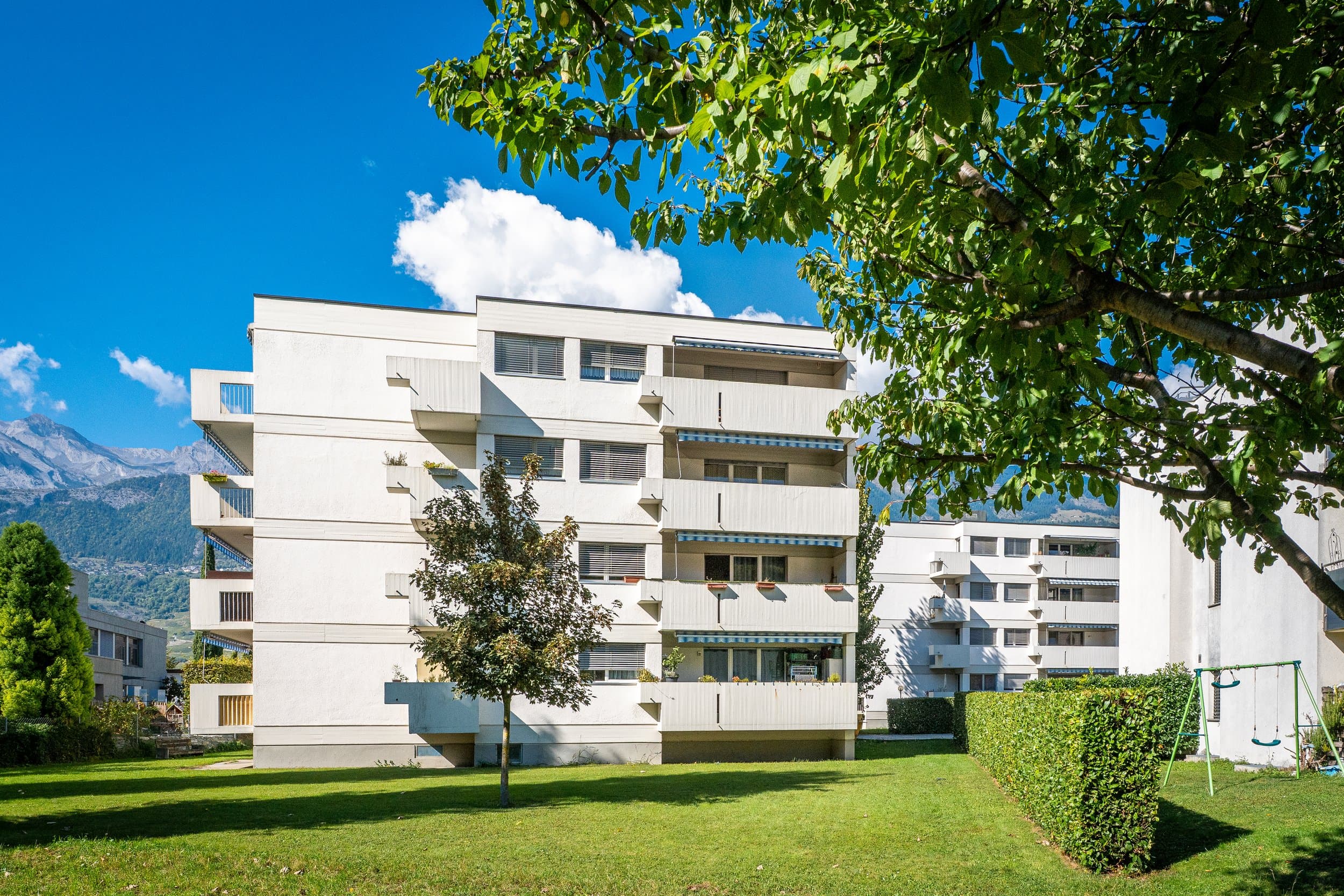 Immeuble locatif blanc à balcons de la Fondation Mon Foyer à Pont-de-la-Morge, Sion, vu depuis une pelouse sous un grand arbre