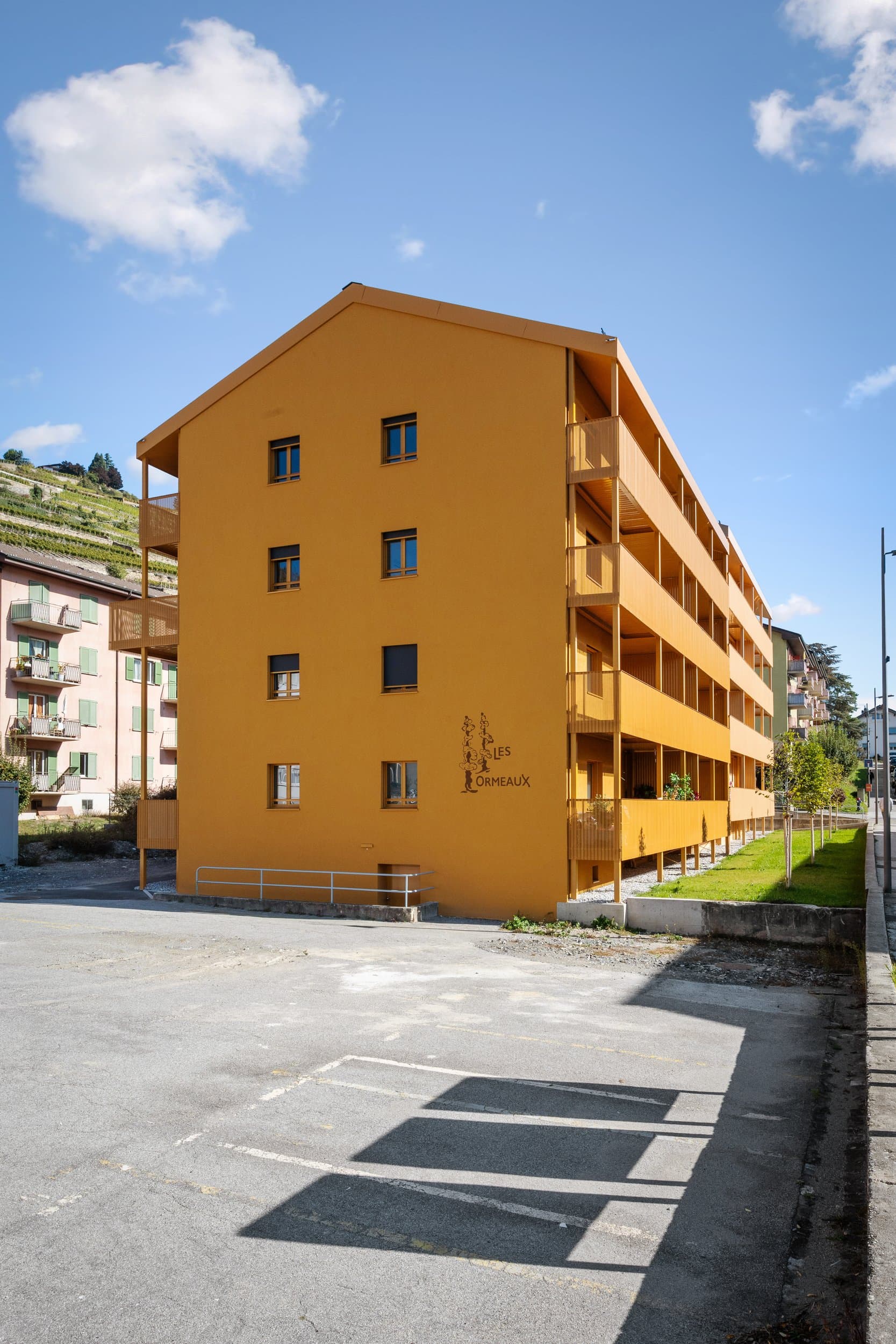 Immeuble aux façades et aux balcons orange, rue des Vignettes à Sion, vu depuis un parking sous un ciel bleu