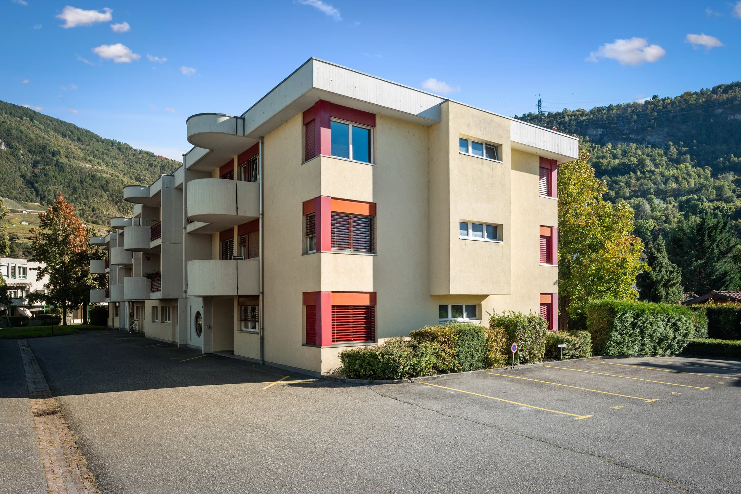 Immeuble crème aux stores rouges et aux balcons arrondis, chemin du Bosquet à Sion, devant son parking sous un ciel bleu