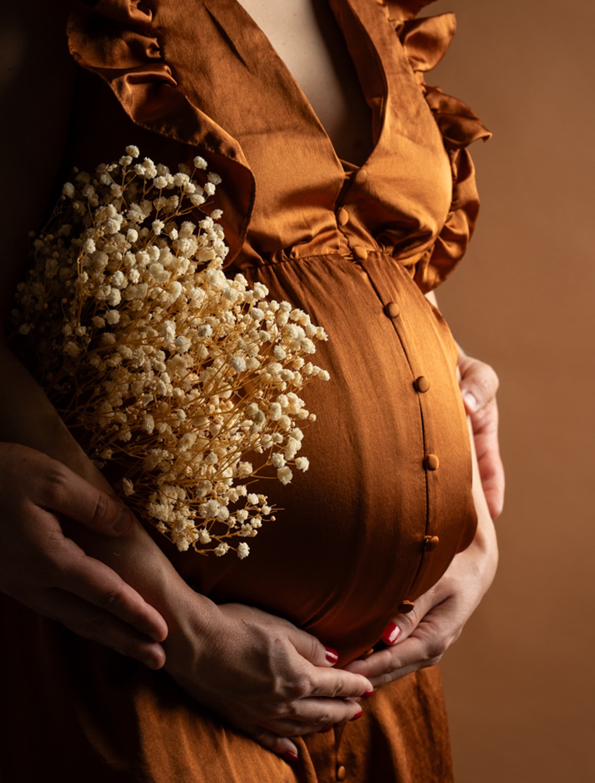 Ventre rond sous une robe en satin rouille, les mains des futurs parents posées dessus, un bouquet de gypsophile