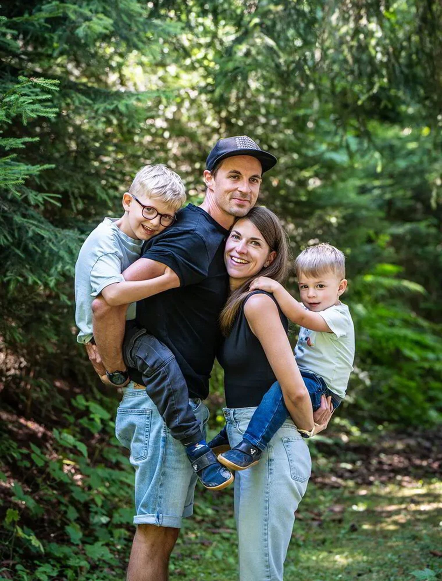 Un papa porte son fils sur le dos et une maman son fils dans les bras, en forêt, au milieu des sapins