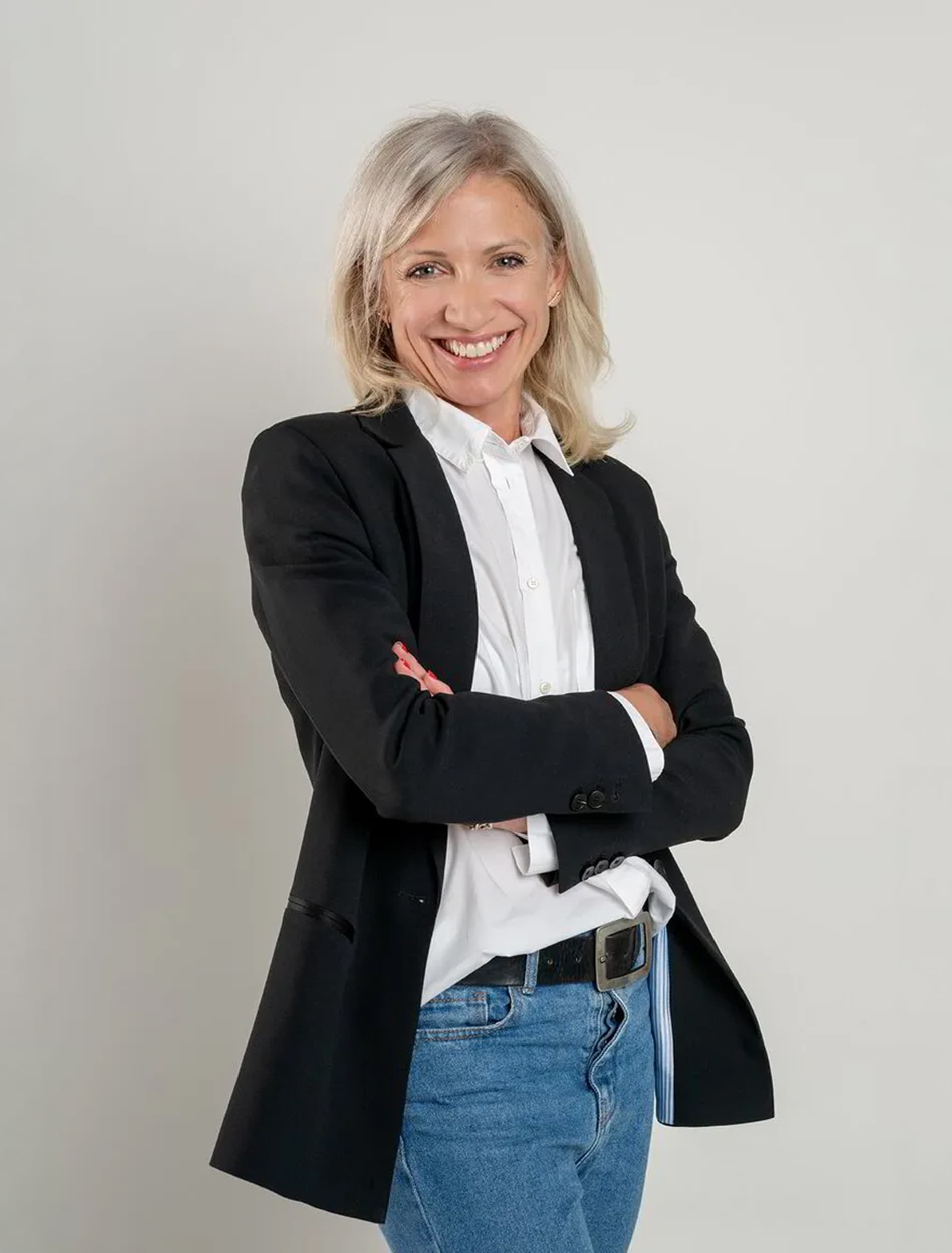 Une femme souriante en blazer noir, bras croisés, sur fond clair