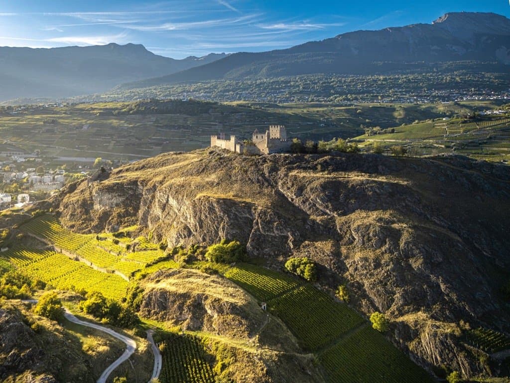 Vue aérienne de la colline de Tourbillon et de son château, entourés de vignes, la plaine du Rhône et les montagnes au loin