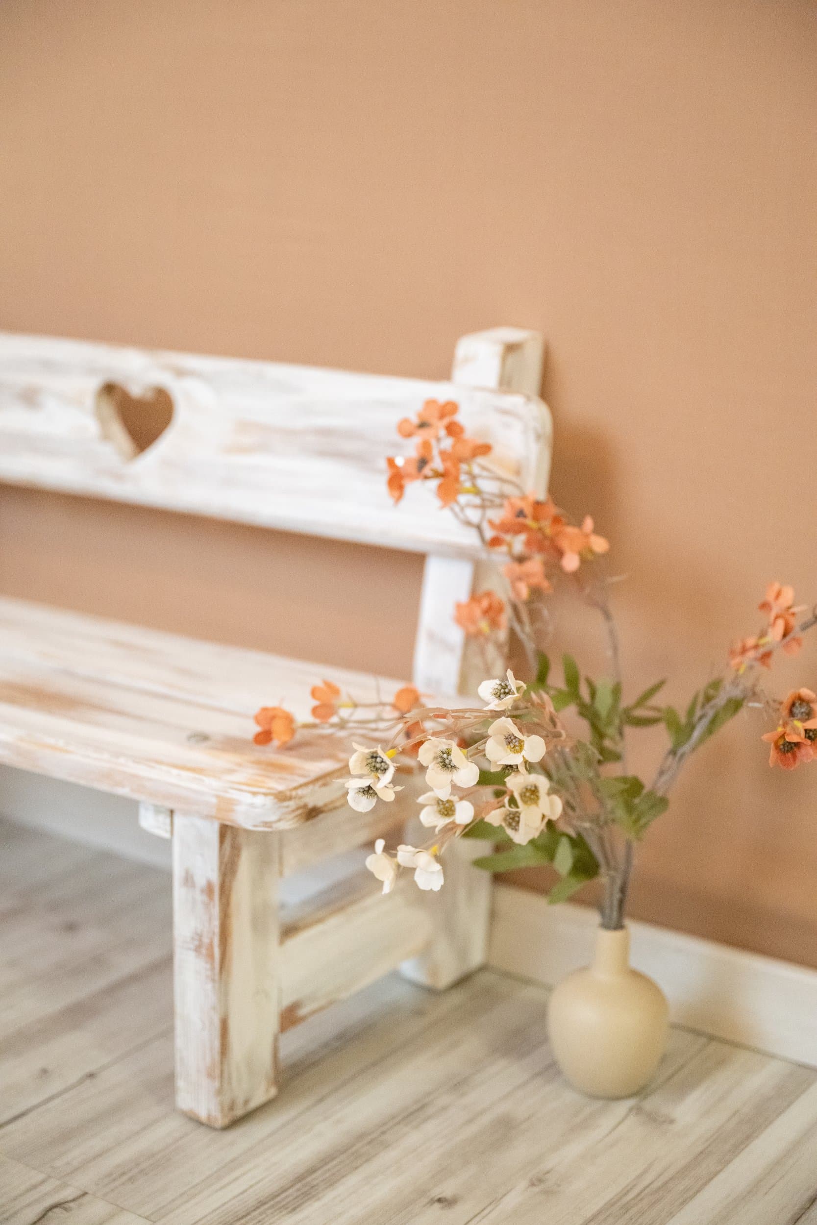Un petit banc en bois blanc patiné, découpé d’un cœur, et une branche de fleurs orangées dans un vase, contre un mur terracotta
