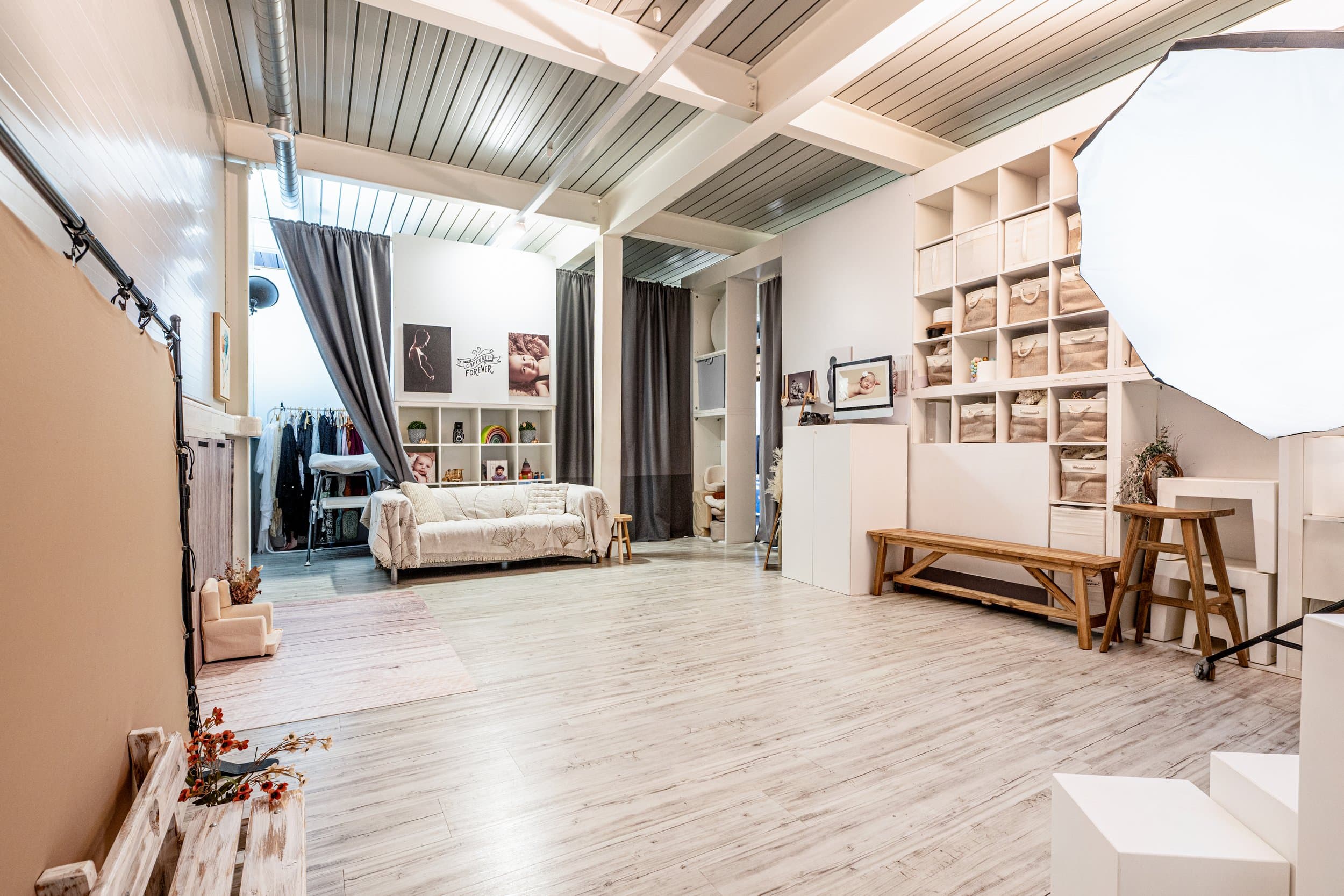 L’Espace principal du studio : un canapé clair, un portant de vêtements, des étagères de paniers, un banc en bois et une grande boîte à lumière, sous un plafond en lames de bois