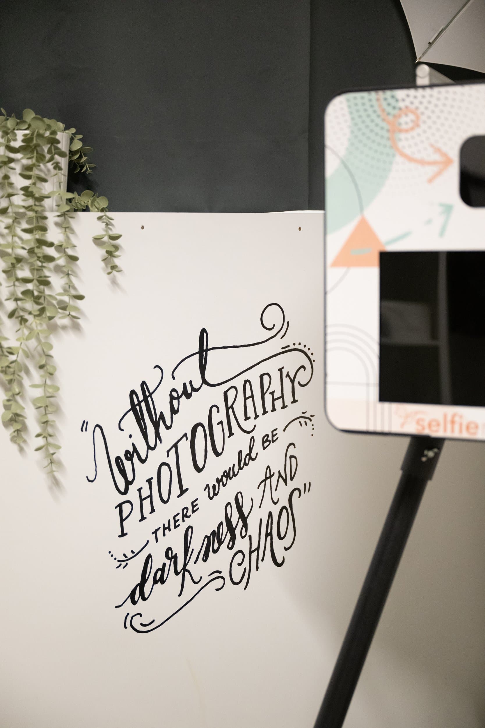 Un mur blanc calligraphié « Without photography there would be darkness and chaos », une plante retombante et un Selfiematon sur son pied