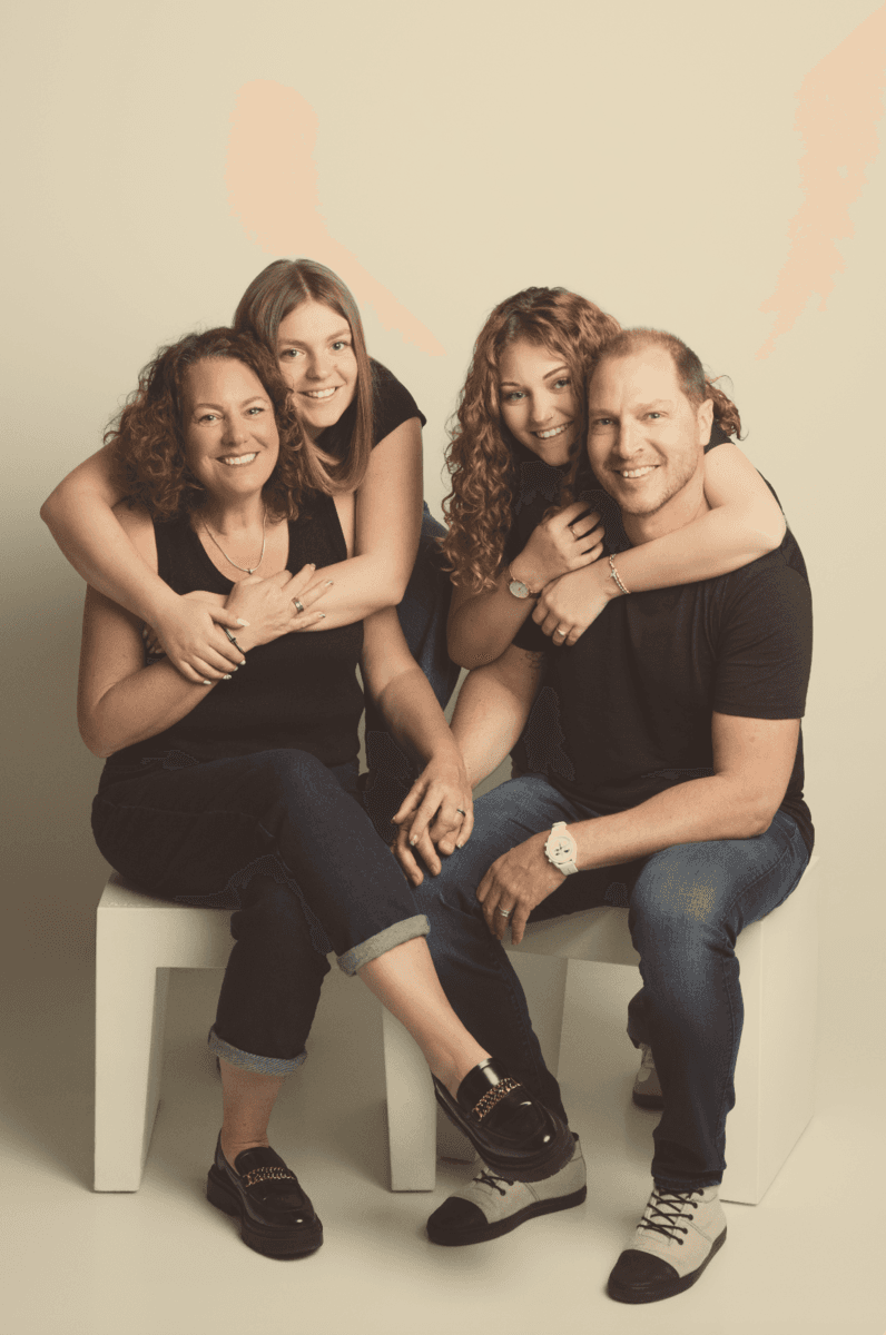 Des parents souriants assis sur des cubes blancs, leurs deux filles les enlacent par derrière, en studio