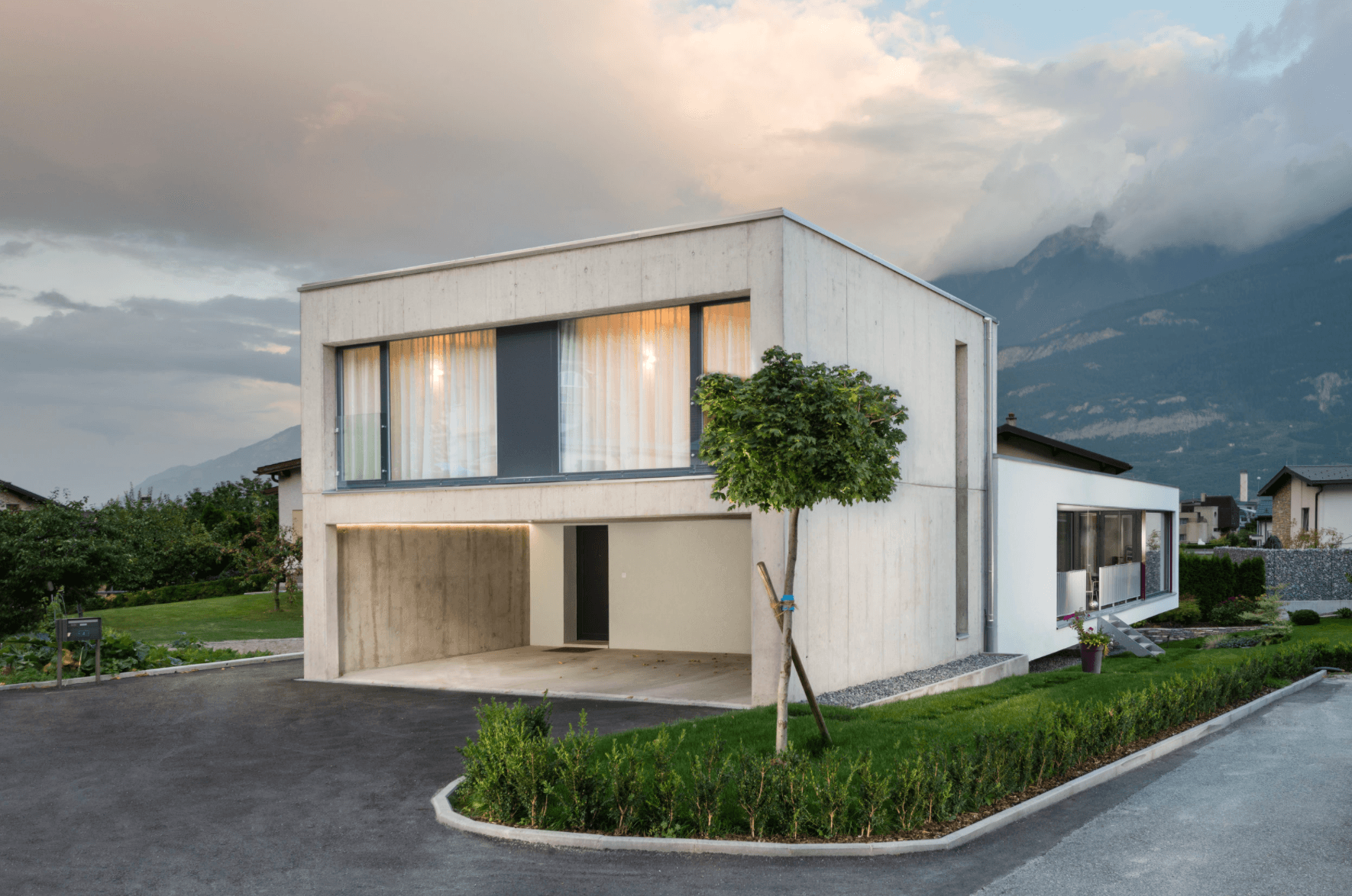 Maison contemporaine en béton clair au bord d’une route, montagnes et nuages en arrière-plan