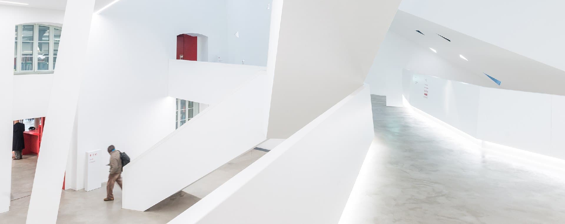 Hall intérieur tout blanc aux parois obliques, une personne monte un escalier