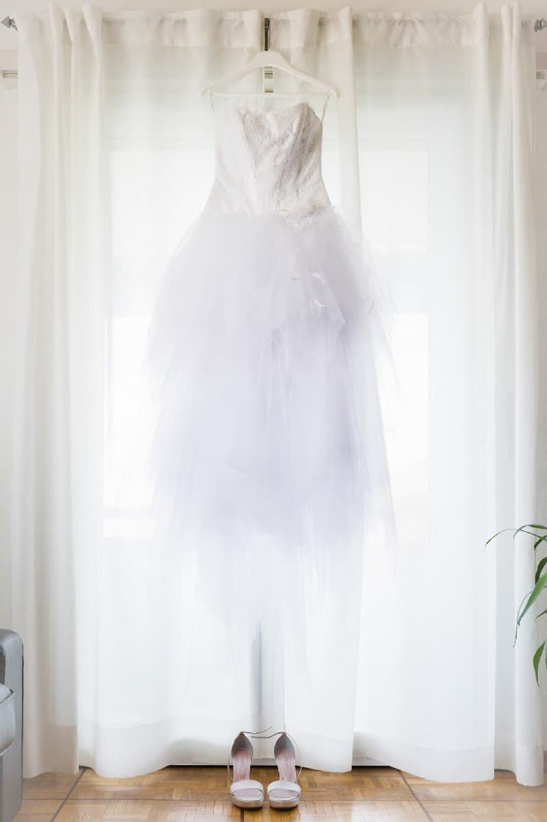 La robe de mariée suspendue devant une fenêtre voilée, les chaussures posées au sol
