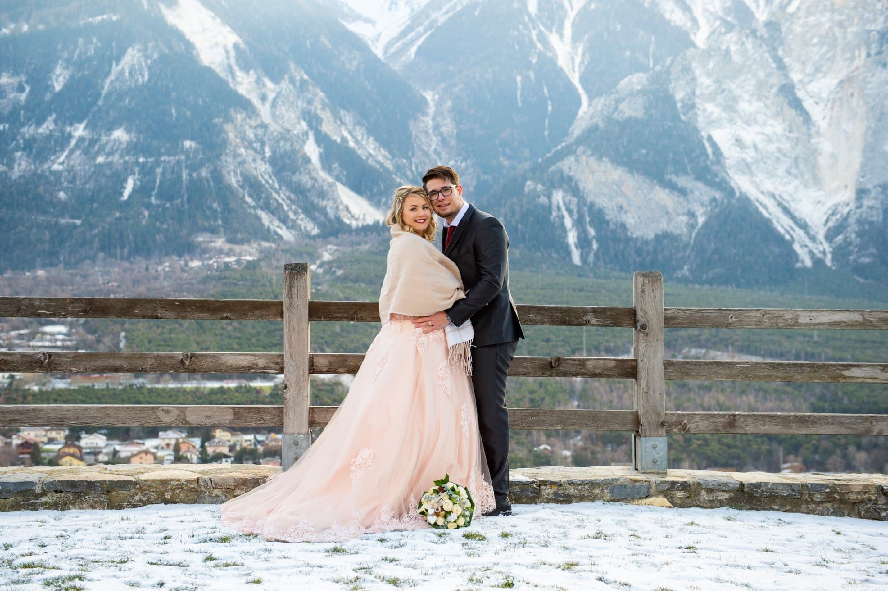 Mariage d’hiver : les mariés devant une barrière en bois, montagnes enneigées derrière eux