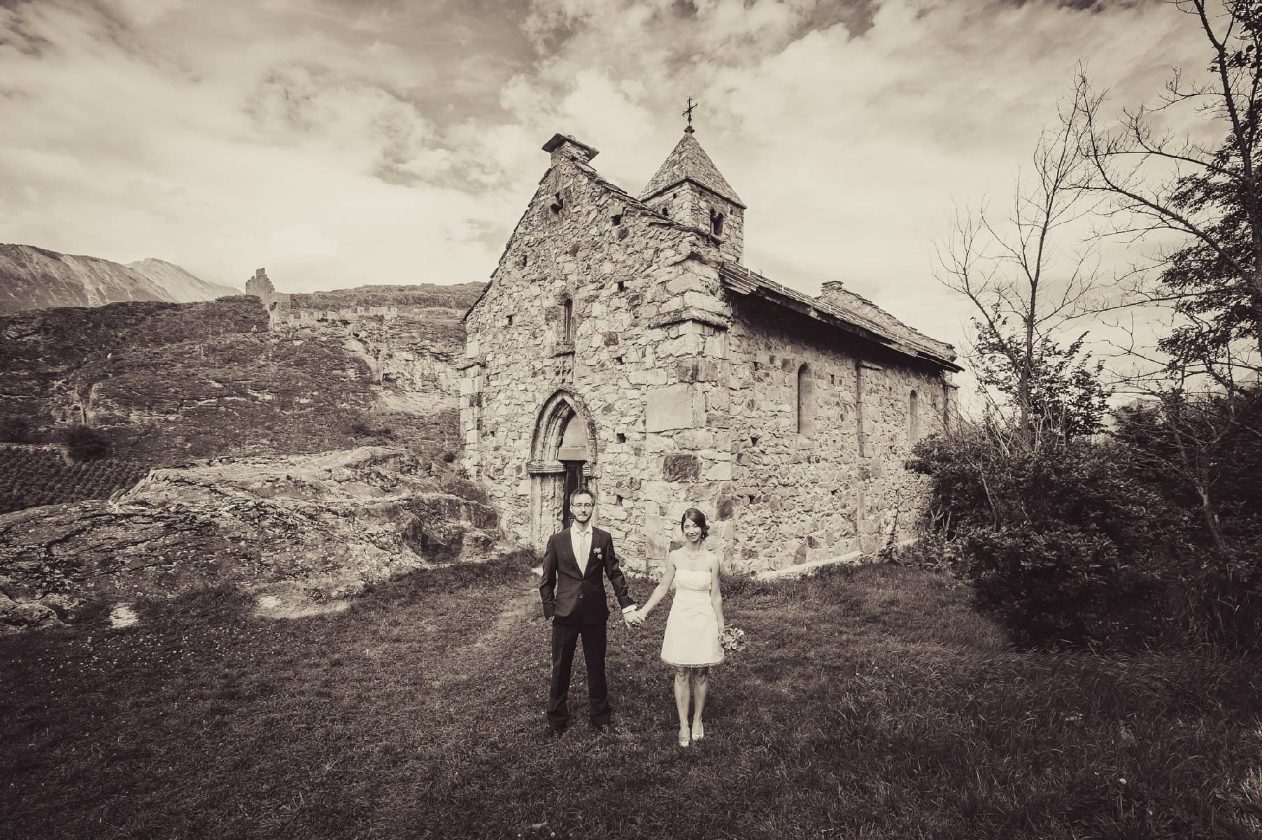 Les mariés main dans la main devant une chapelle en pierre, photo en tons sépia