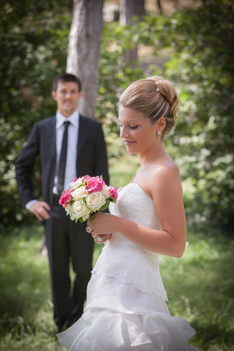 La mariée regarde son bouquet, le marié flou derrière elle, sous les arbres