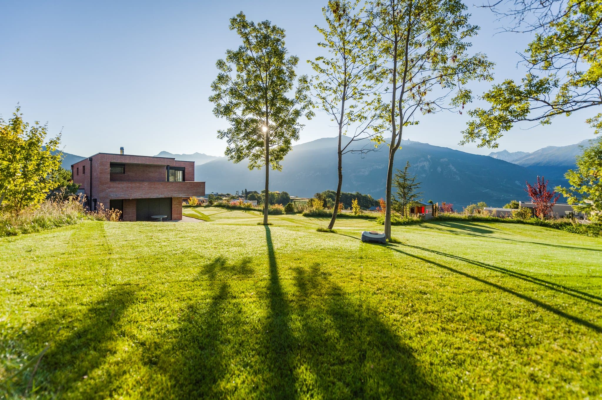 Maison en brique au bout d’une grande pelouse, arbres et montagnes au soleil