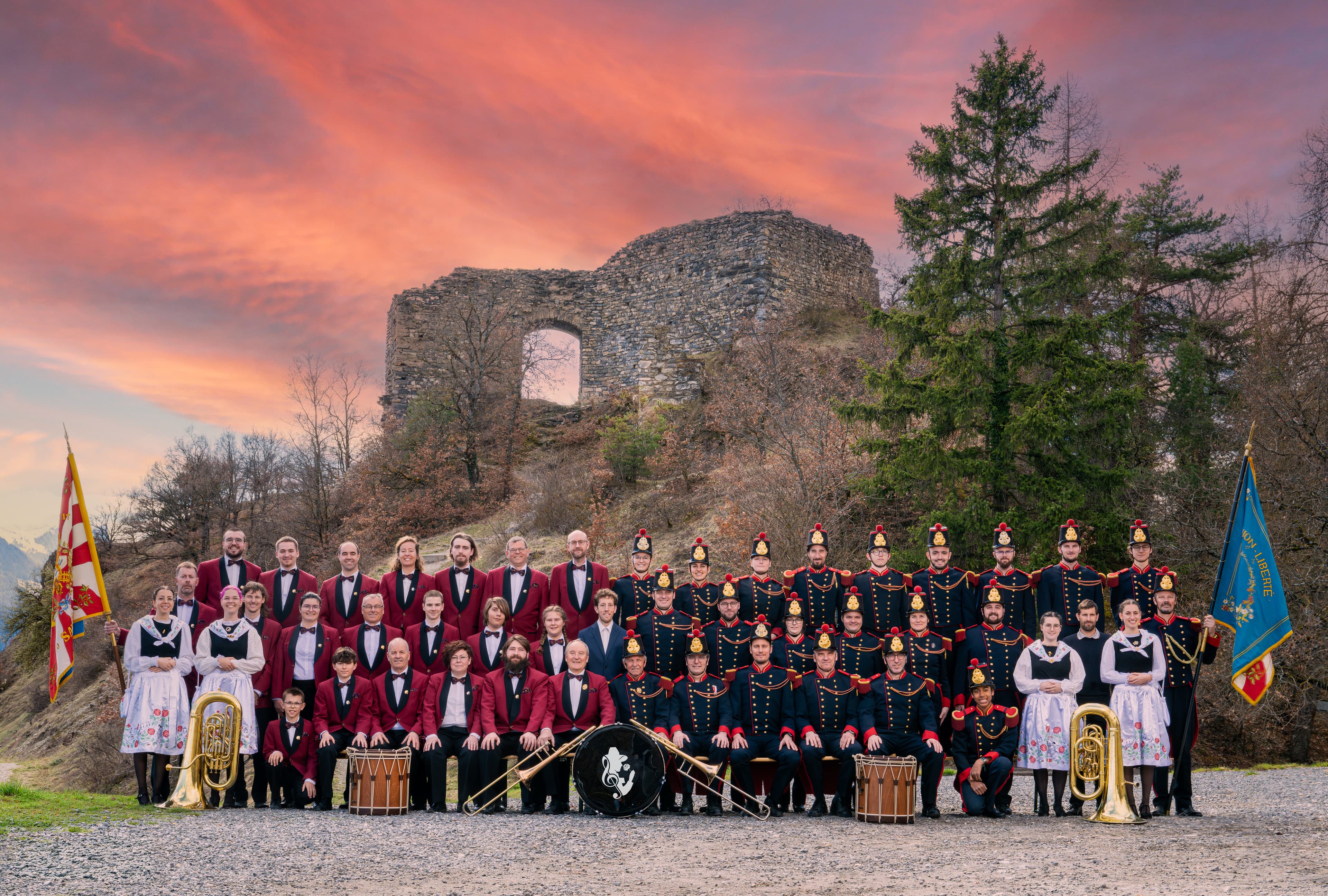 Fanfare en uniforme rouge et noir, avec des personnes en costume traditionnel, posée devant une ruine de château au coucher du soleil