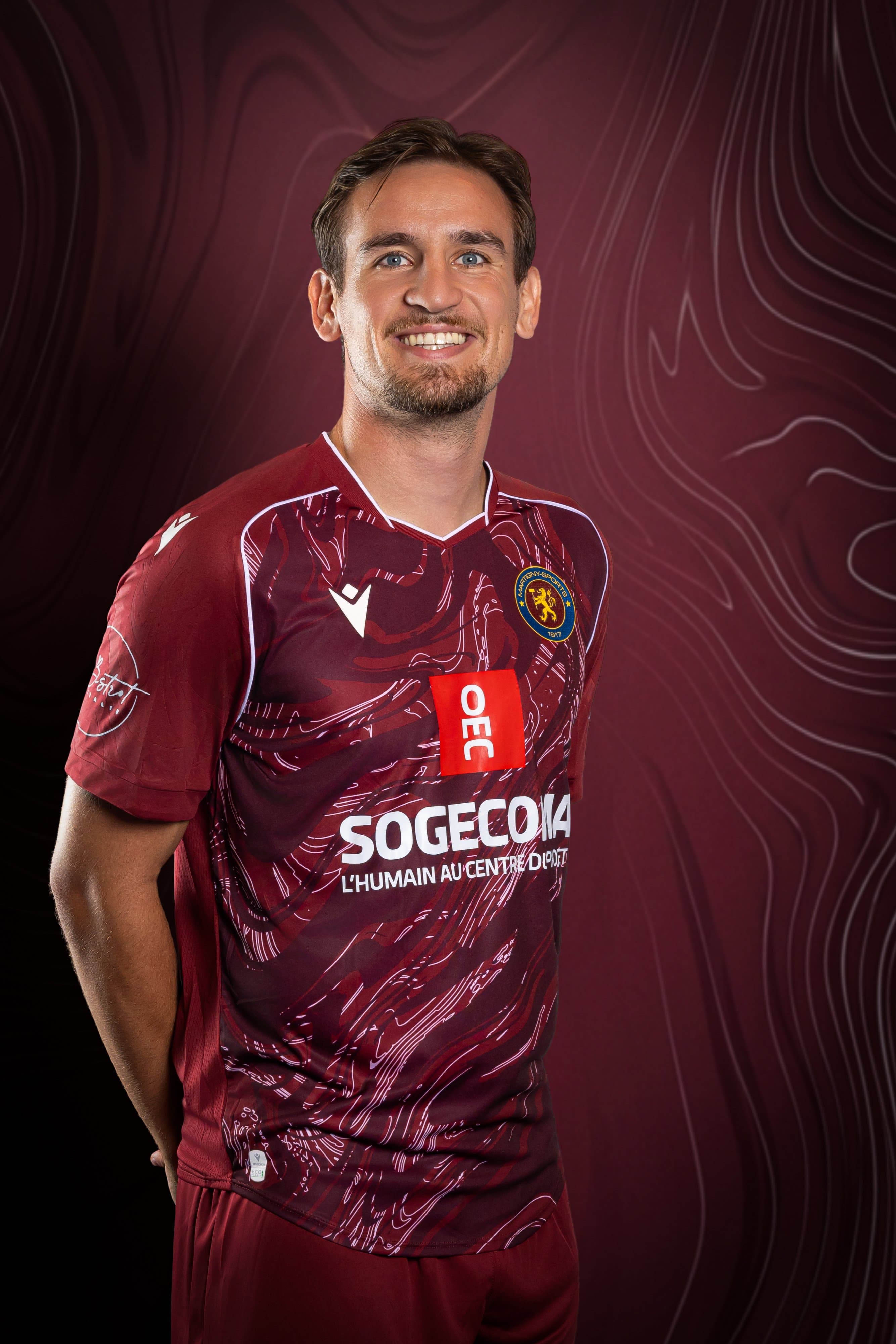 Portrait d’un joueur souriant du Martigny-Sports en maillot bordeaux, sur fond bordeaux