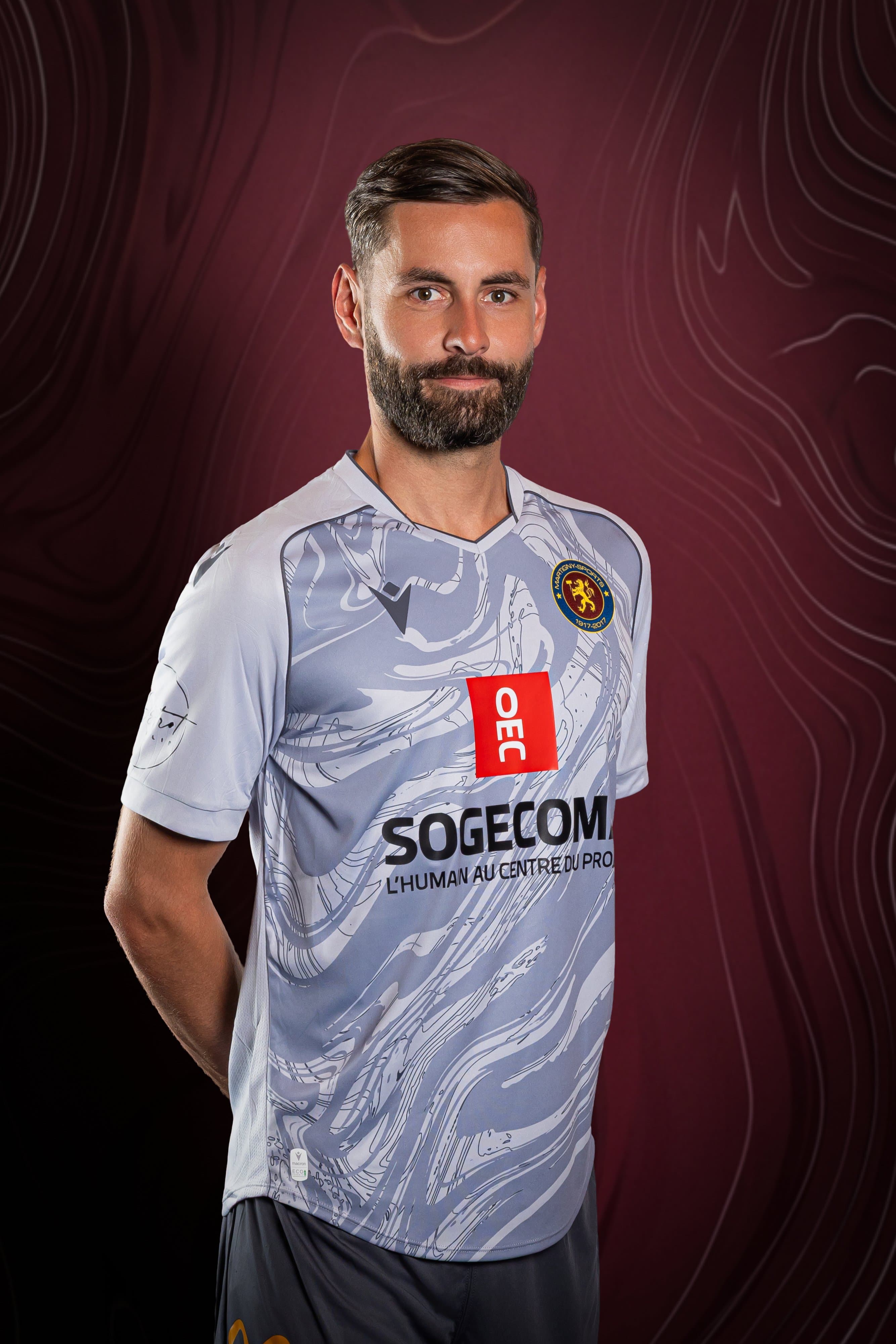 Portrait d’un joueur barbu du Martigny-Sports en maillot gris, sur fond bordeaux