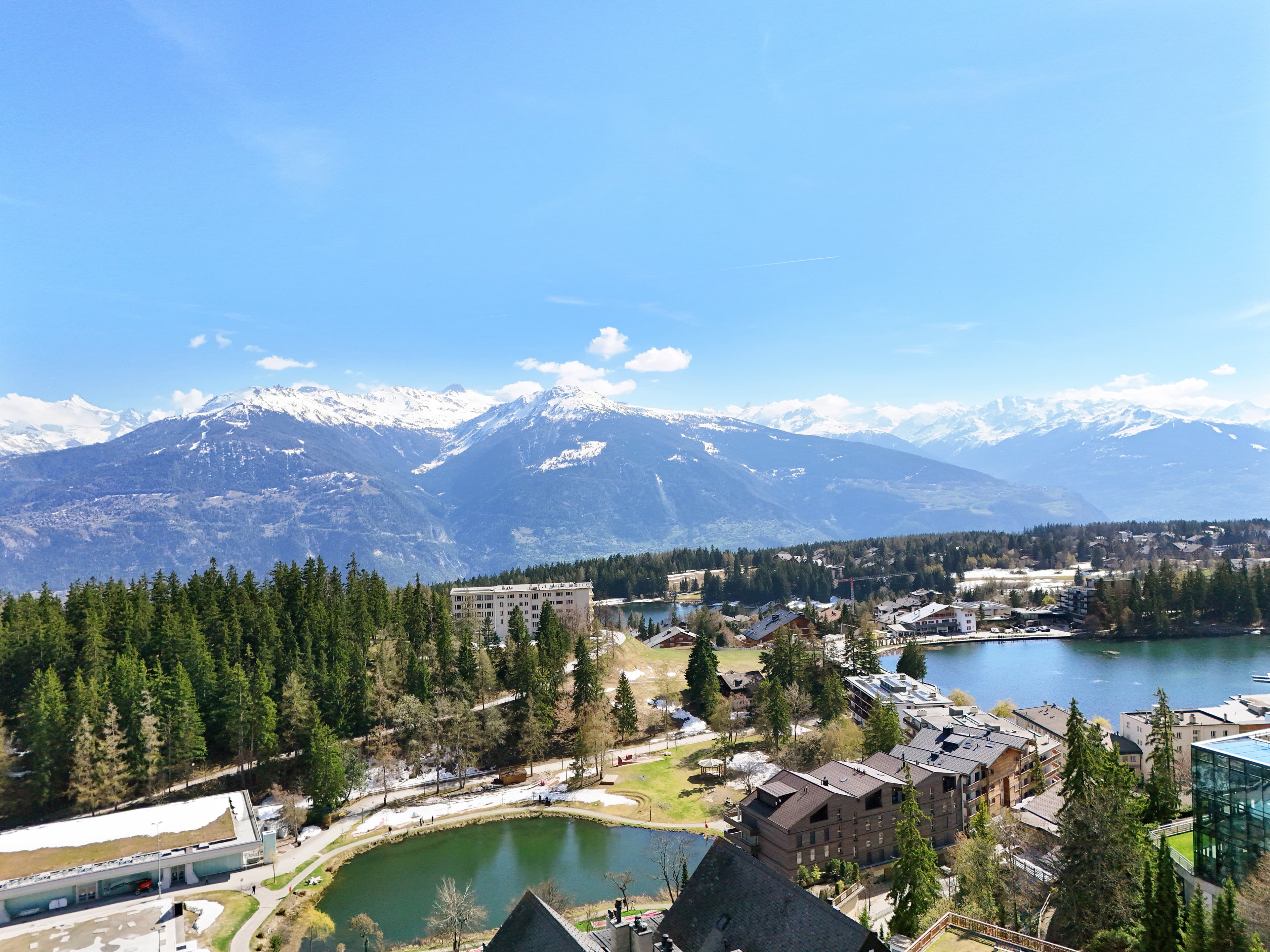 Vue aérienne de Crans-Montana : forêts, lacs et chaîne des Alpes enneigée sous le ciel bleu