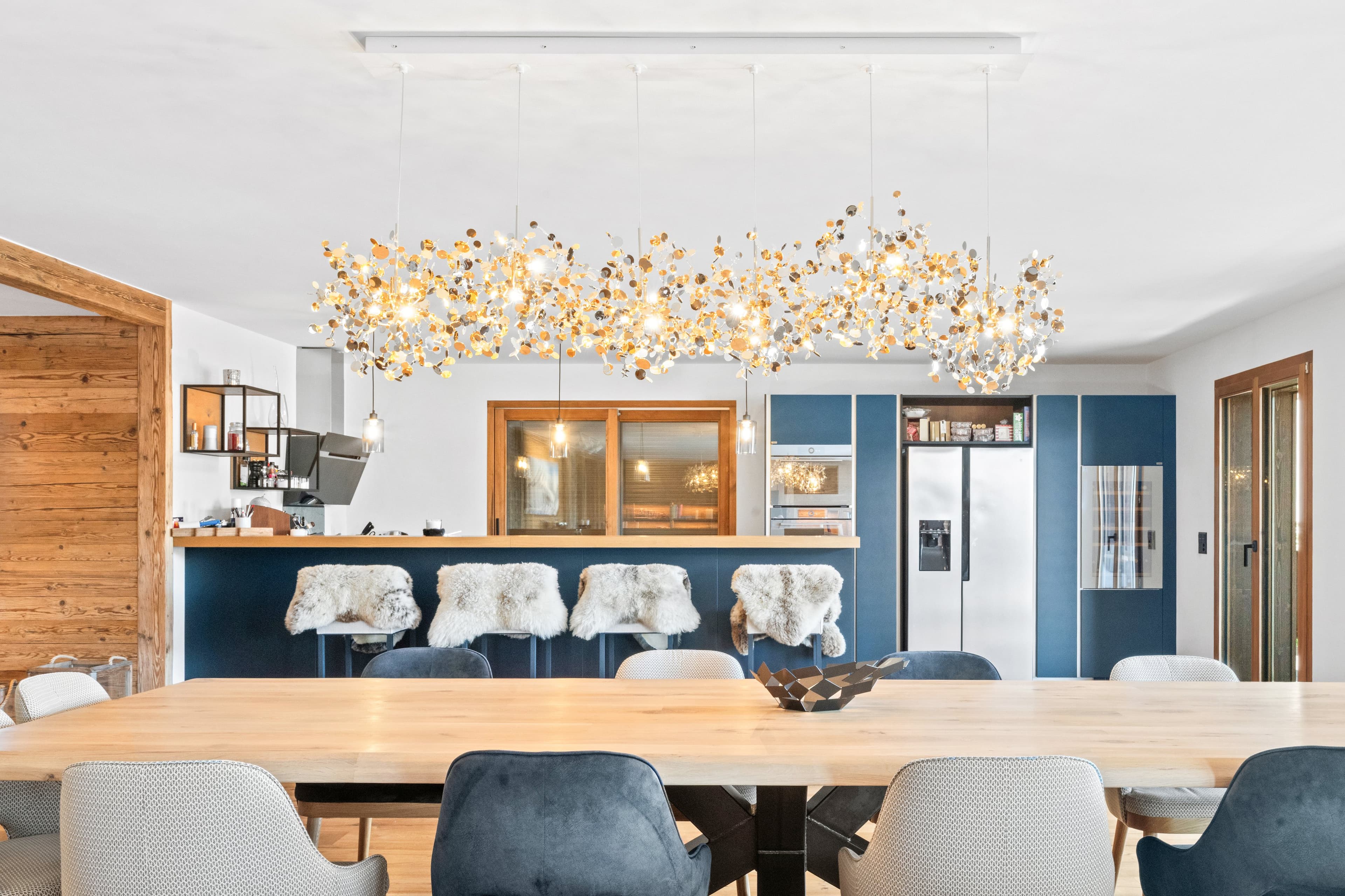 Salle à manger sous un grand lustre doré, longue table en bois face à la cuisine bleue