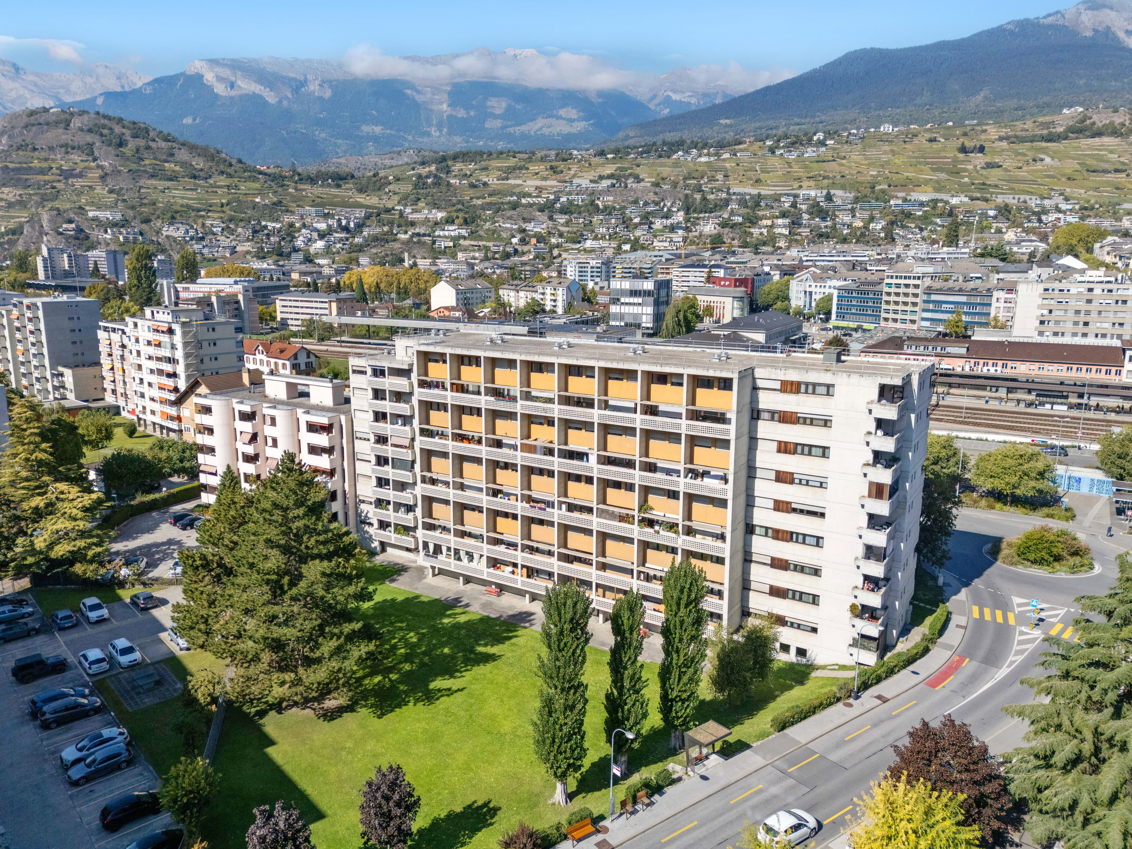 Vue aérienne d’un immeuble de logements à balcons jaunes, son parc arboré et le quartier, montagnes en arrière-plan
