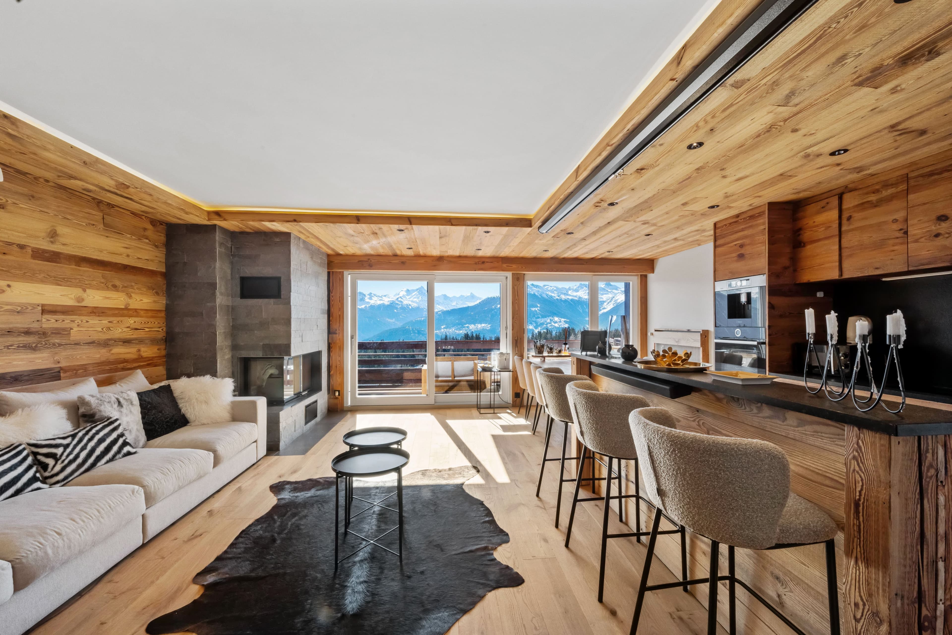Séjour de chalet tout en bois : canapé blanc, cheminée, bar de cuisine et baies vitrées sur les Alpes enneigées