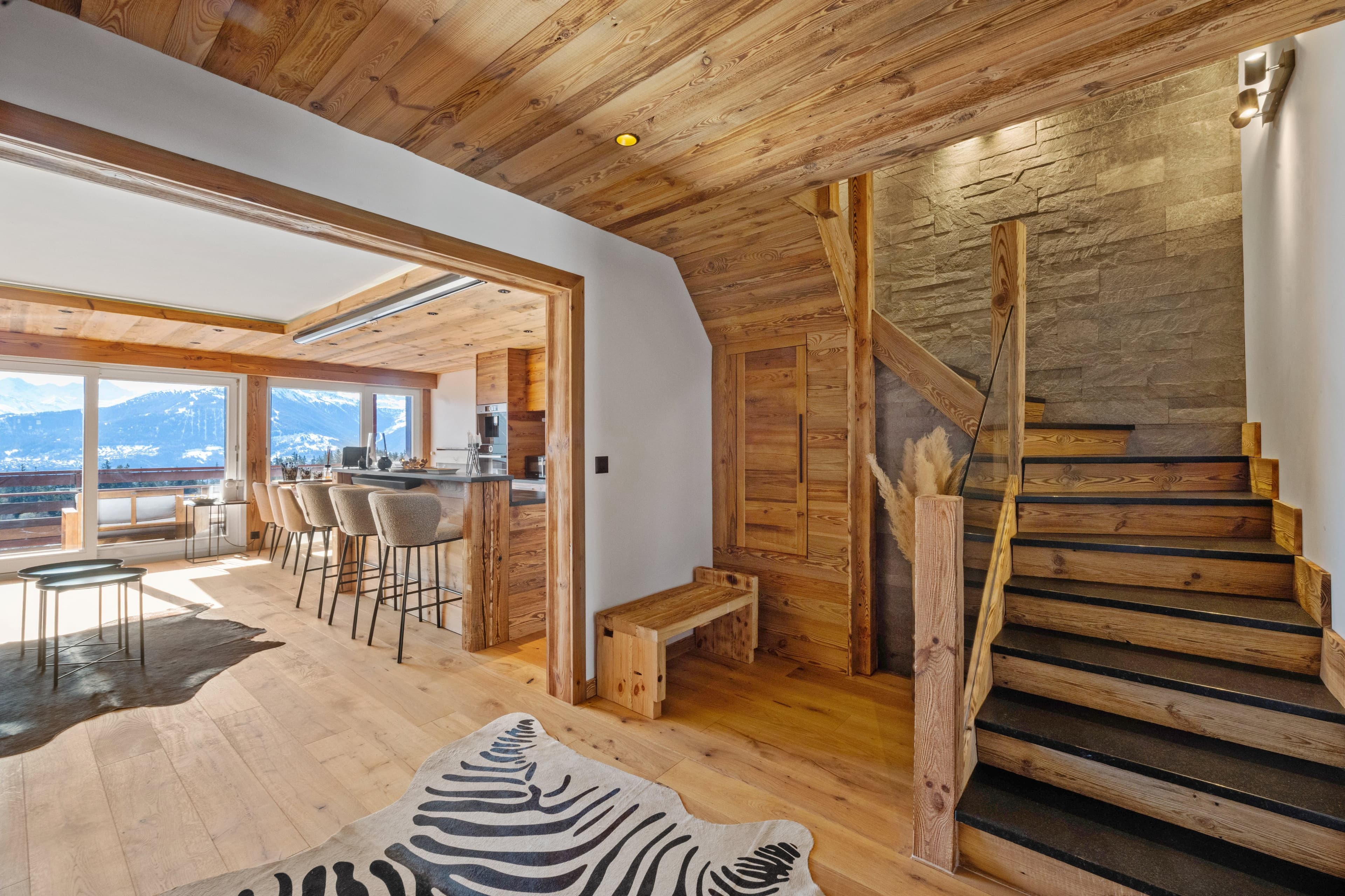 Séjour de chalet avec escalier en bois et pierre, bar de cuisine et fenêtres sur les montagnes