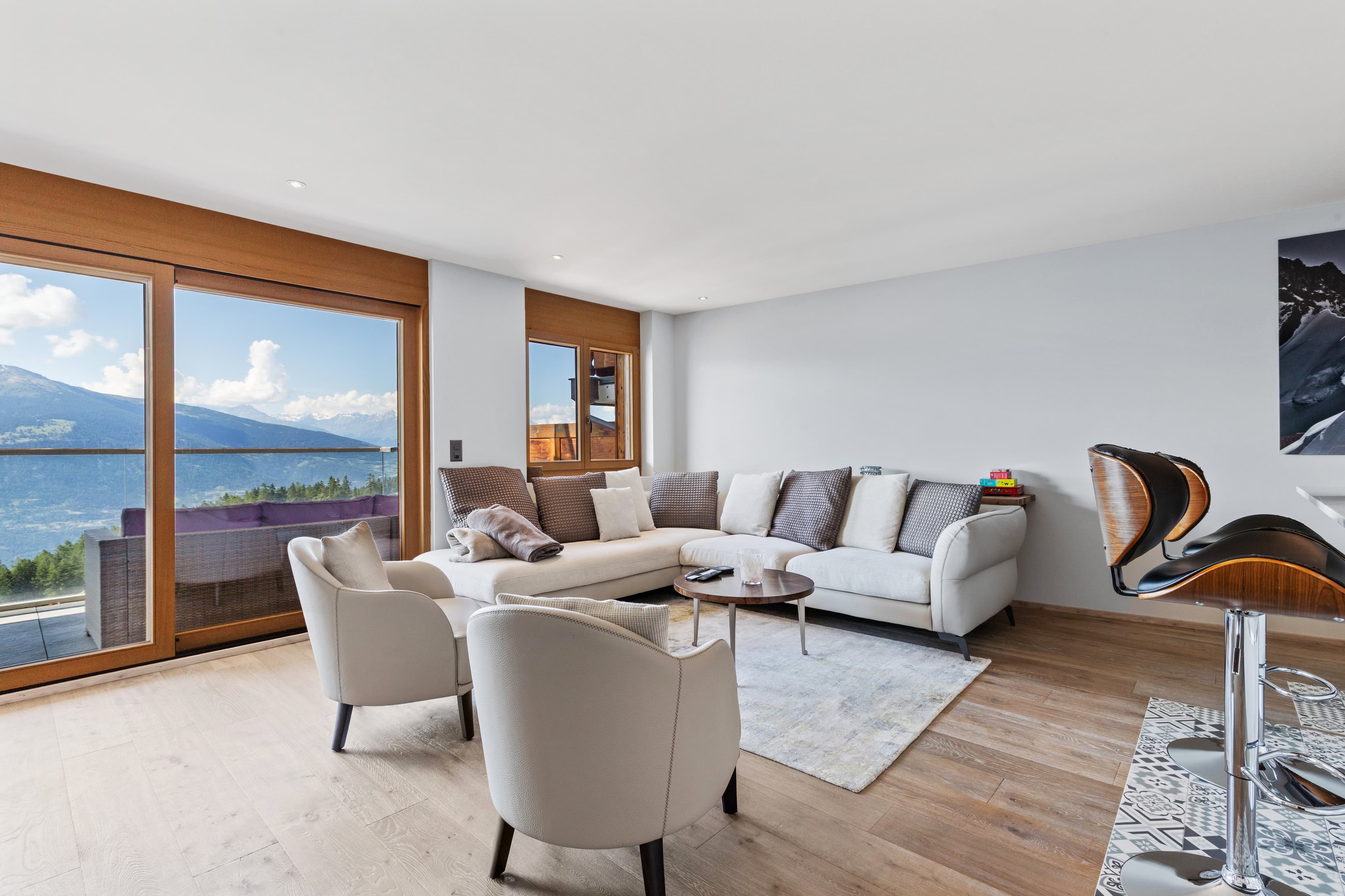 Salon lumineux d’un appartement en station, canapé d’angle et fauteuils blancs devant une baie vitrée ouverte sur la vallée