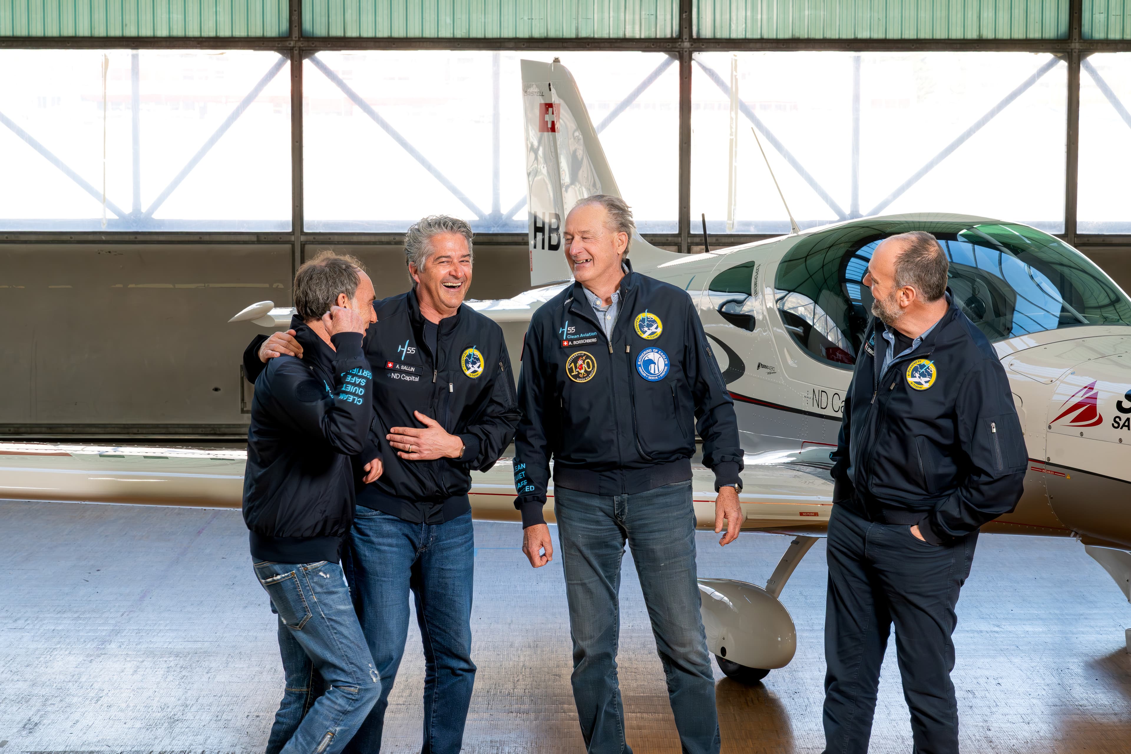 Quatre pilotes en blouson qui rient ensemble devant un petit avion, dans un hangar