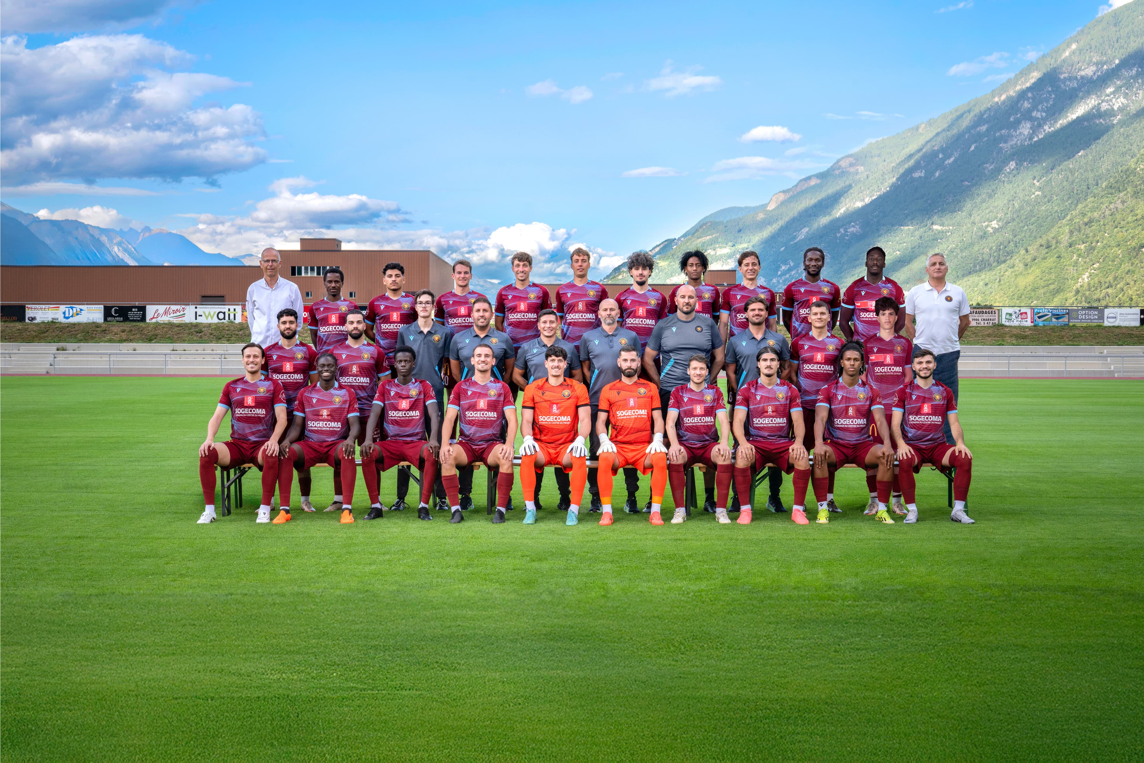 Équipe de football en maillots bordeaux sur deux rangs, devant les tribunes d’un stade et les montagnes