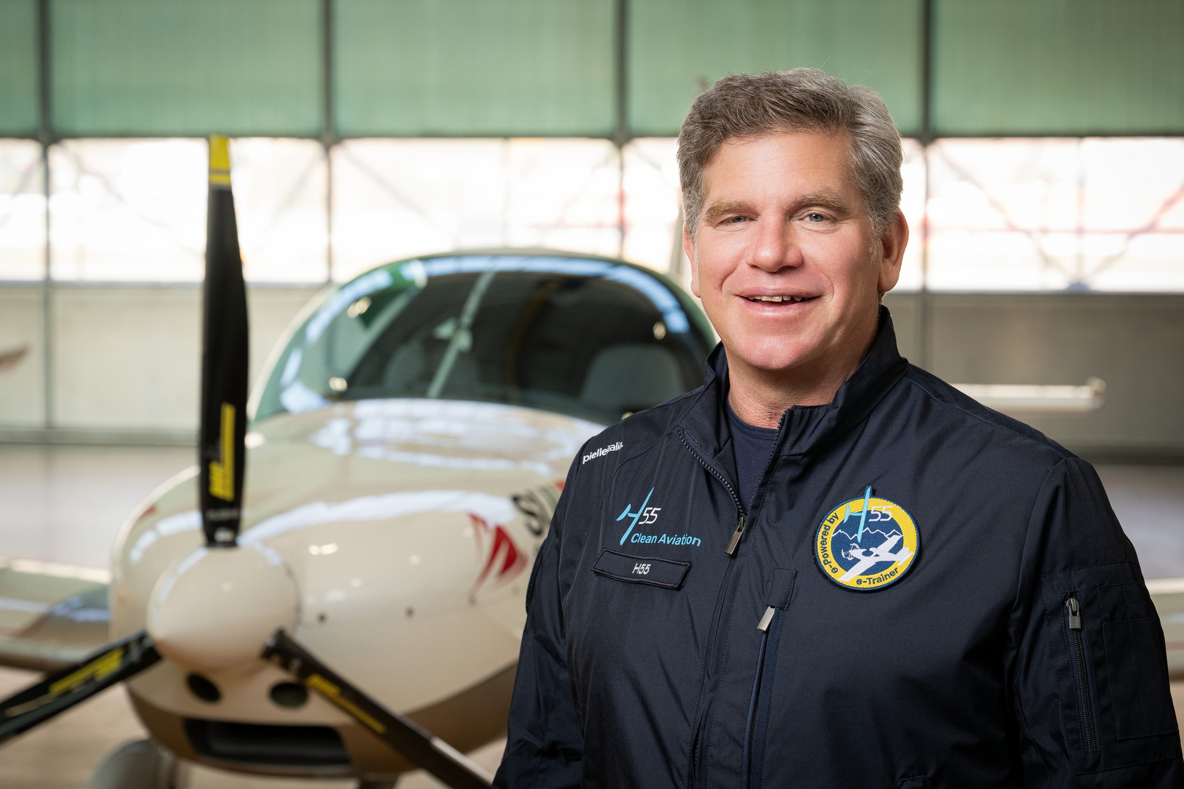 Homme souriant en blouson d’aviateur H55, devant l’hélice d’un avion dans un hangar